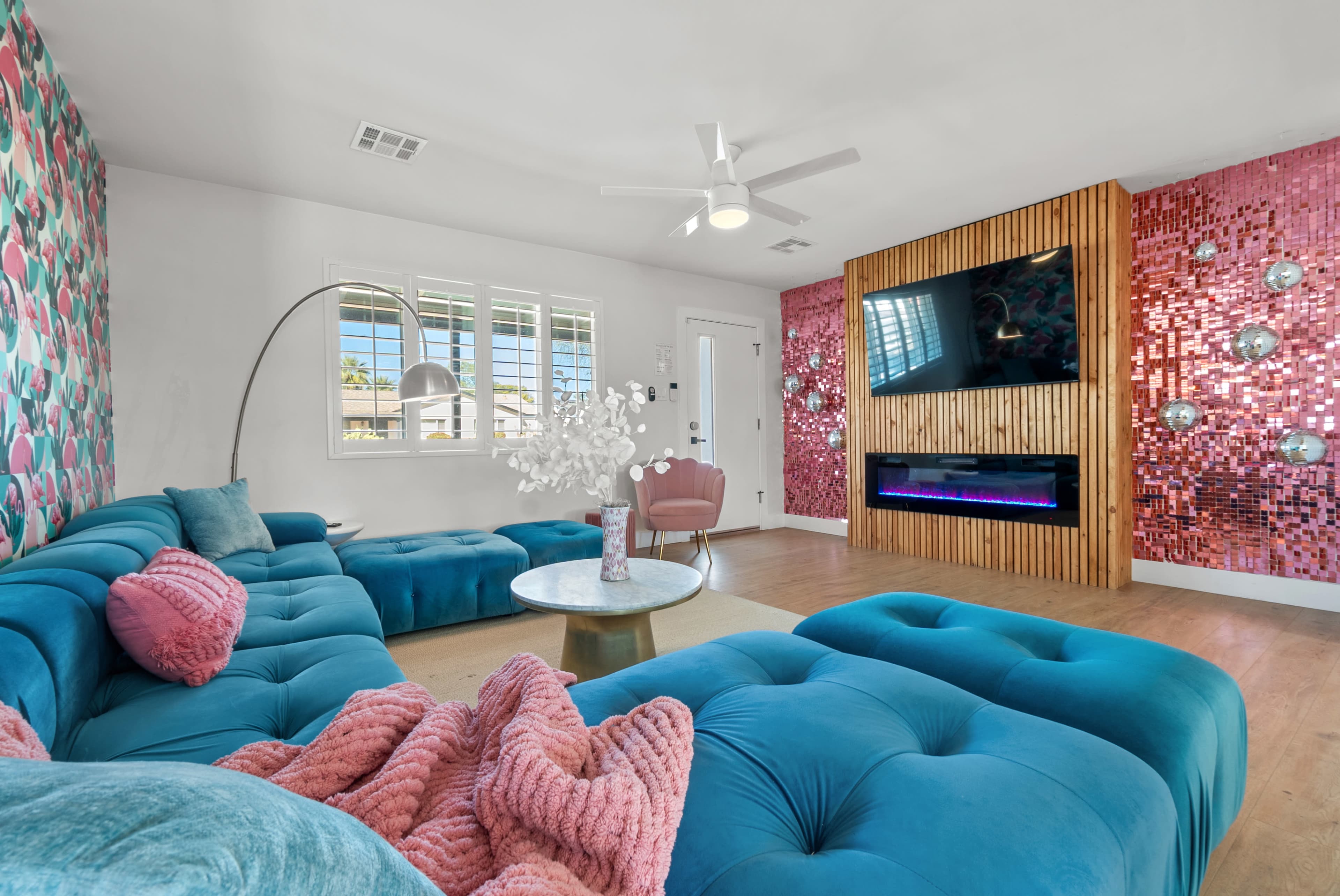 The living room features a large turquoise sectional sofa with a round glass table, a wall with geometric patterns, and a modern fireplace beneath a wall-mounted television.