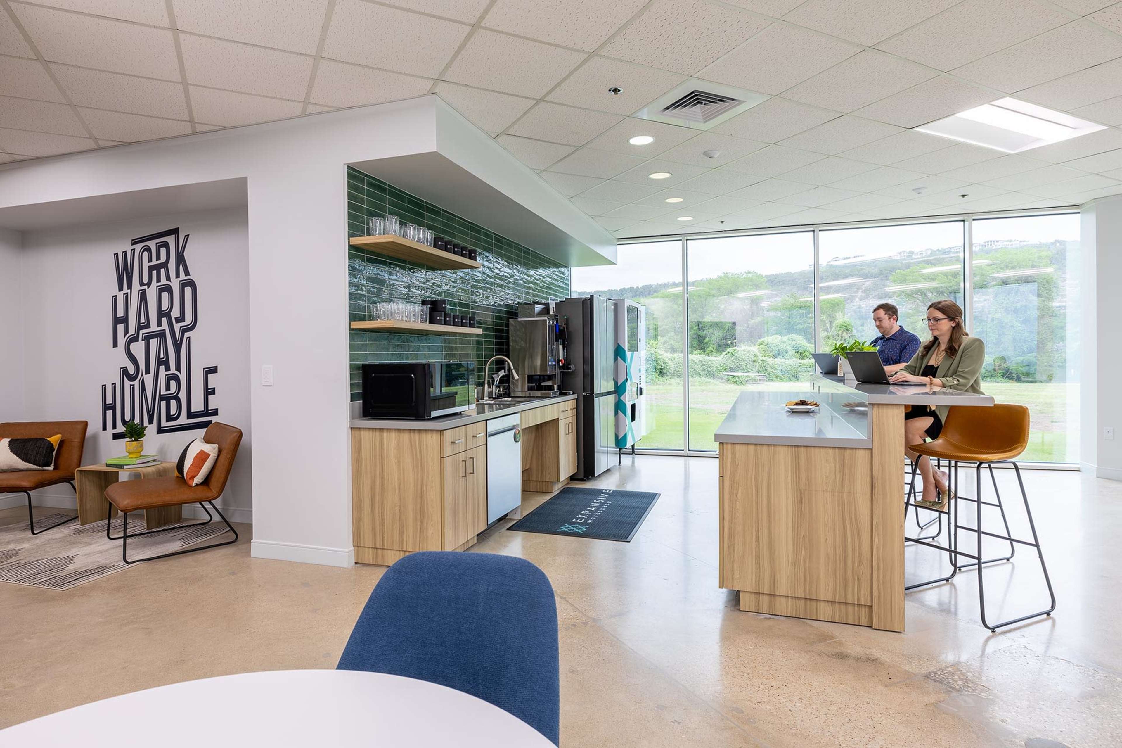 The image features a modern office kitchenette with a large window, where two individuals are engaged in work while sitting at a counter and table, surrounded by stylish furniture and wall art.