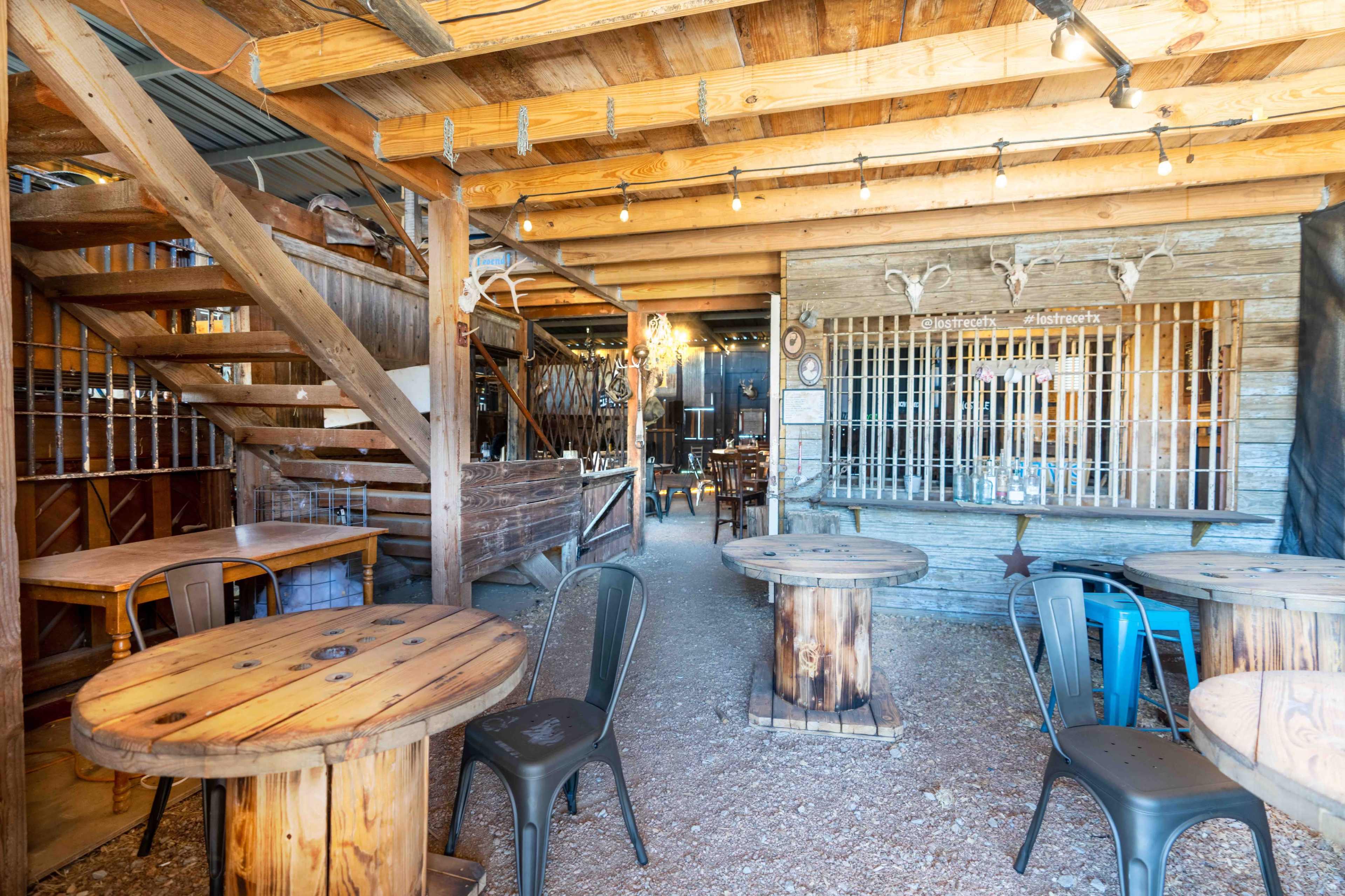 The scene shows a rustic wooden interior of a bar or gathering space with tables, chairs, and a staircase leading to an upper level.