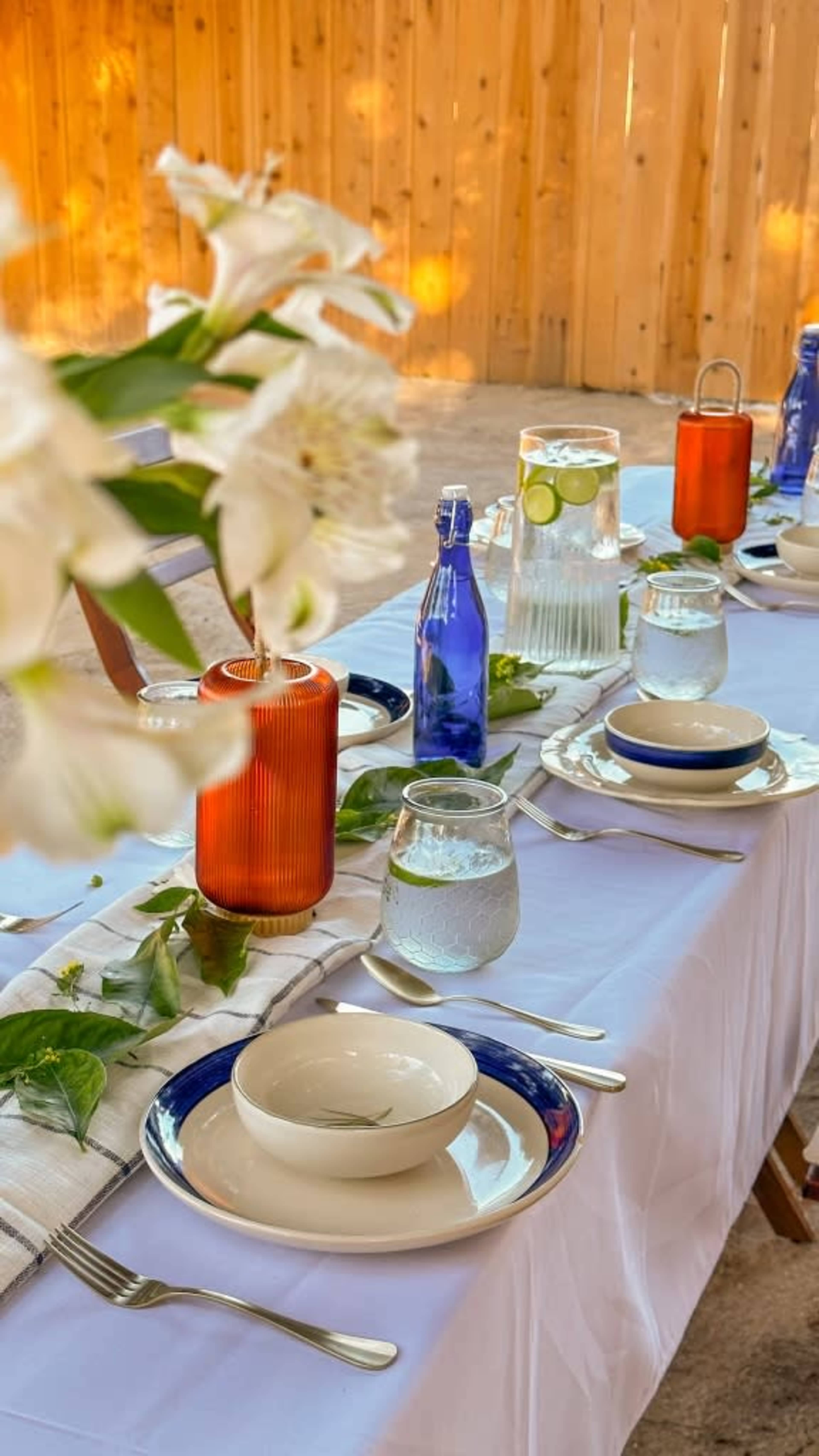 A long outdoor dining table is set with plates, glasses, and decorative vases, surrounded by wooden fencing and greenery.
