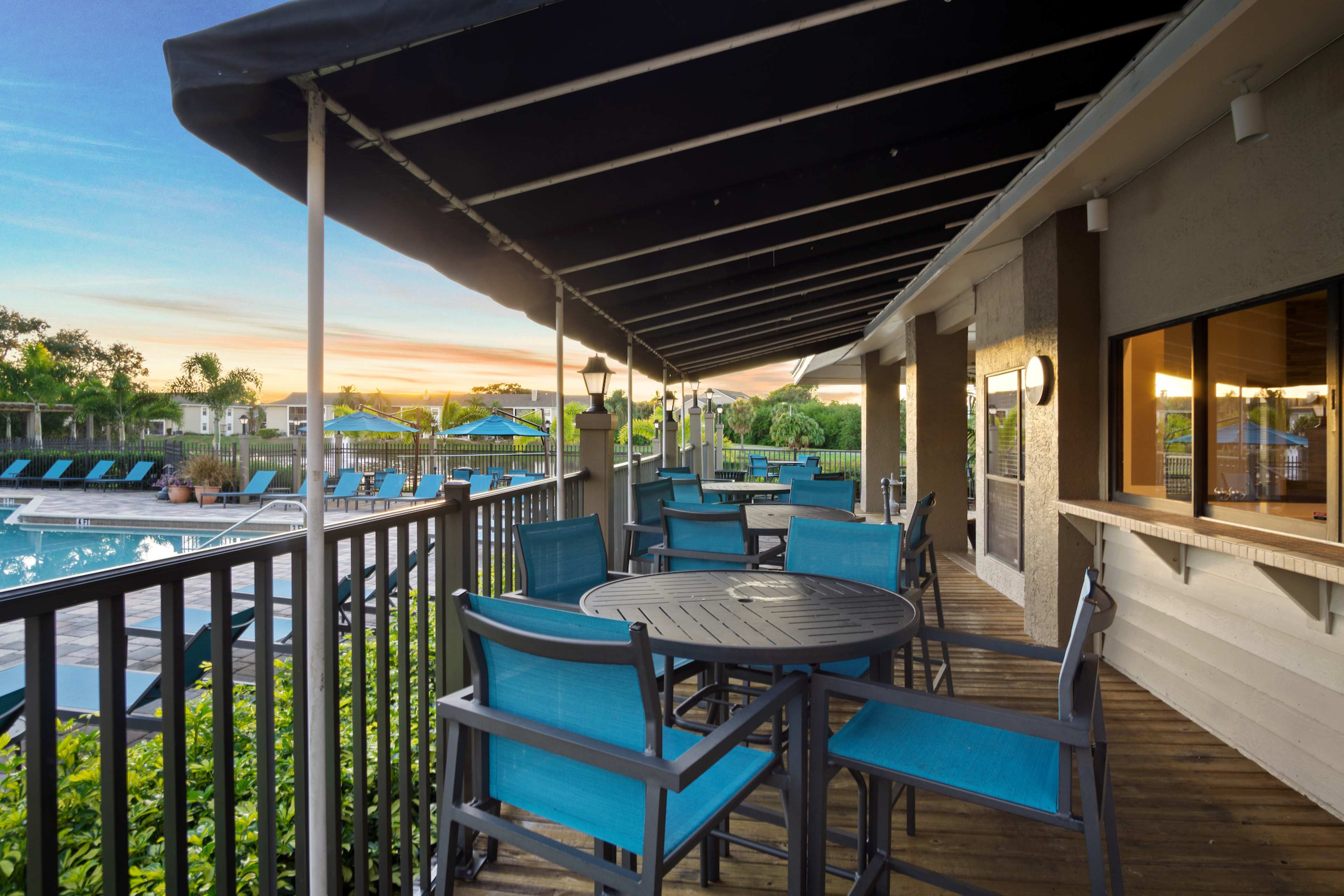 The image shows a patio area with turquoise chairs and tables overlooking a swimming pool at sunset.