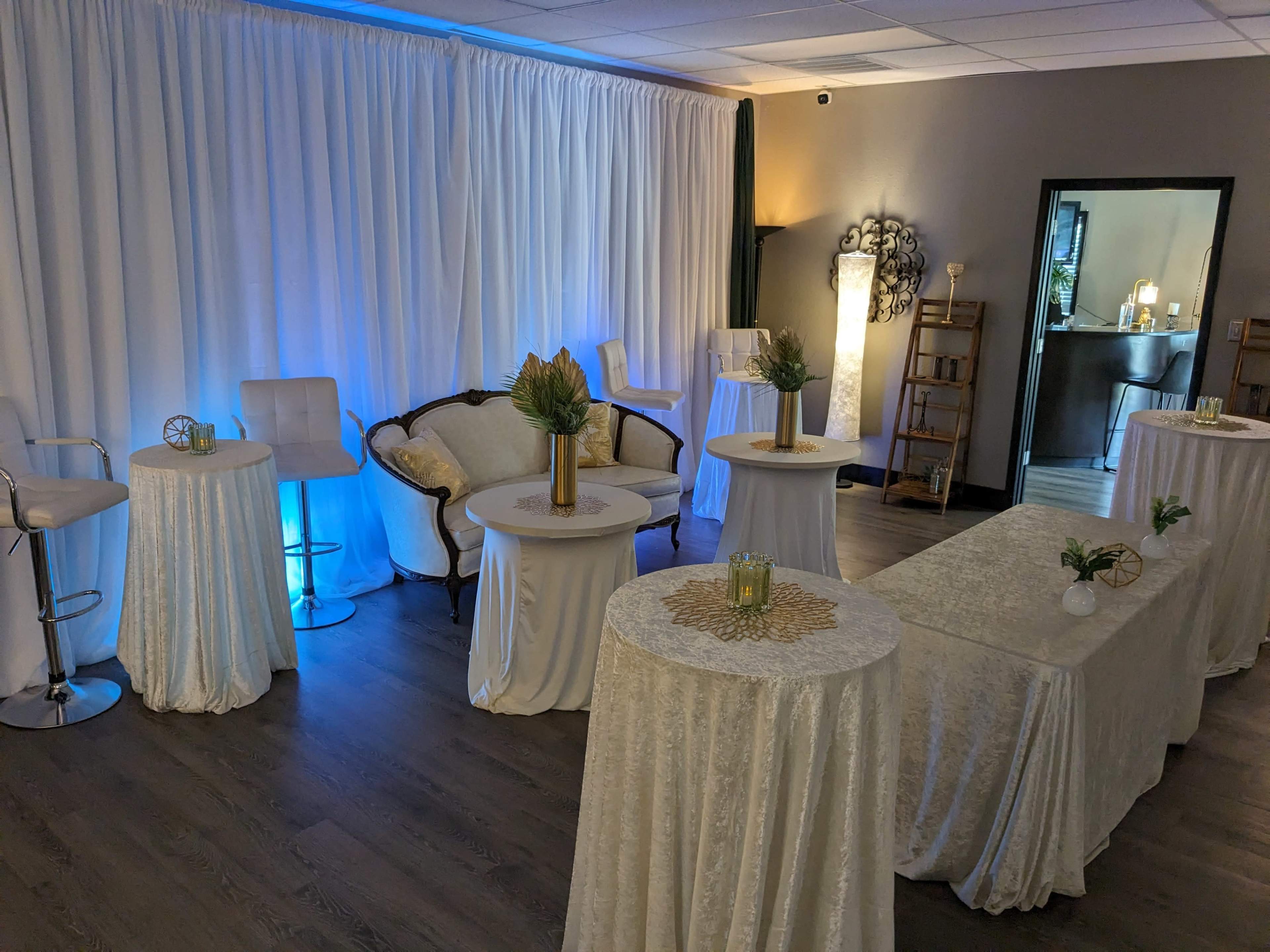 The image features a decorated event space with a white backdrop, several round tables covered in white cloth, a vintage sofa, and various decorative elements.