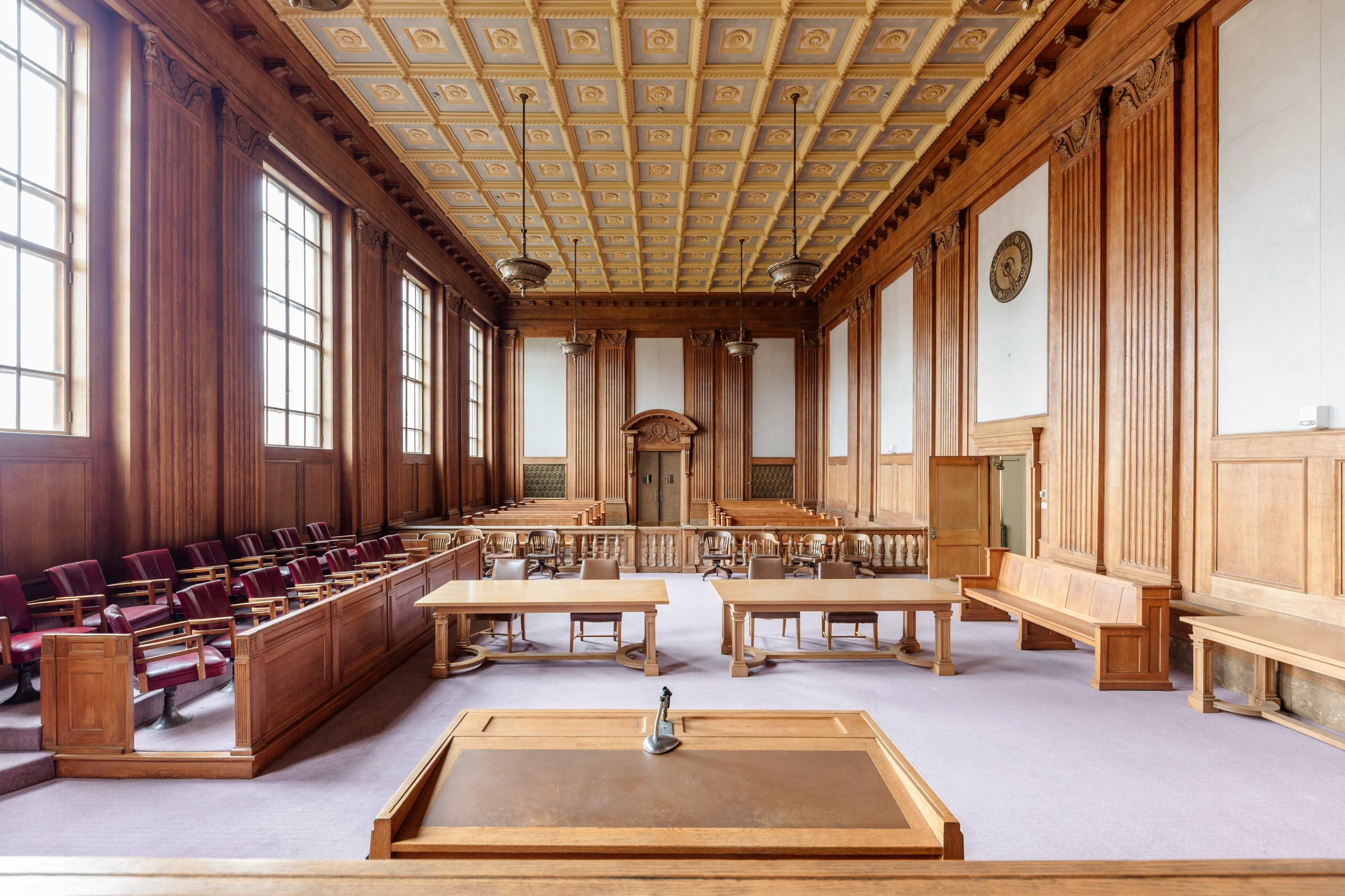 The image shows a spacious courtroom with wooden paneling, large windows, and several tables arranged for legal proceedings.