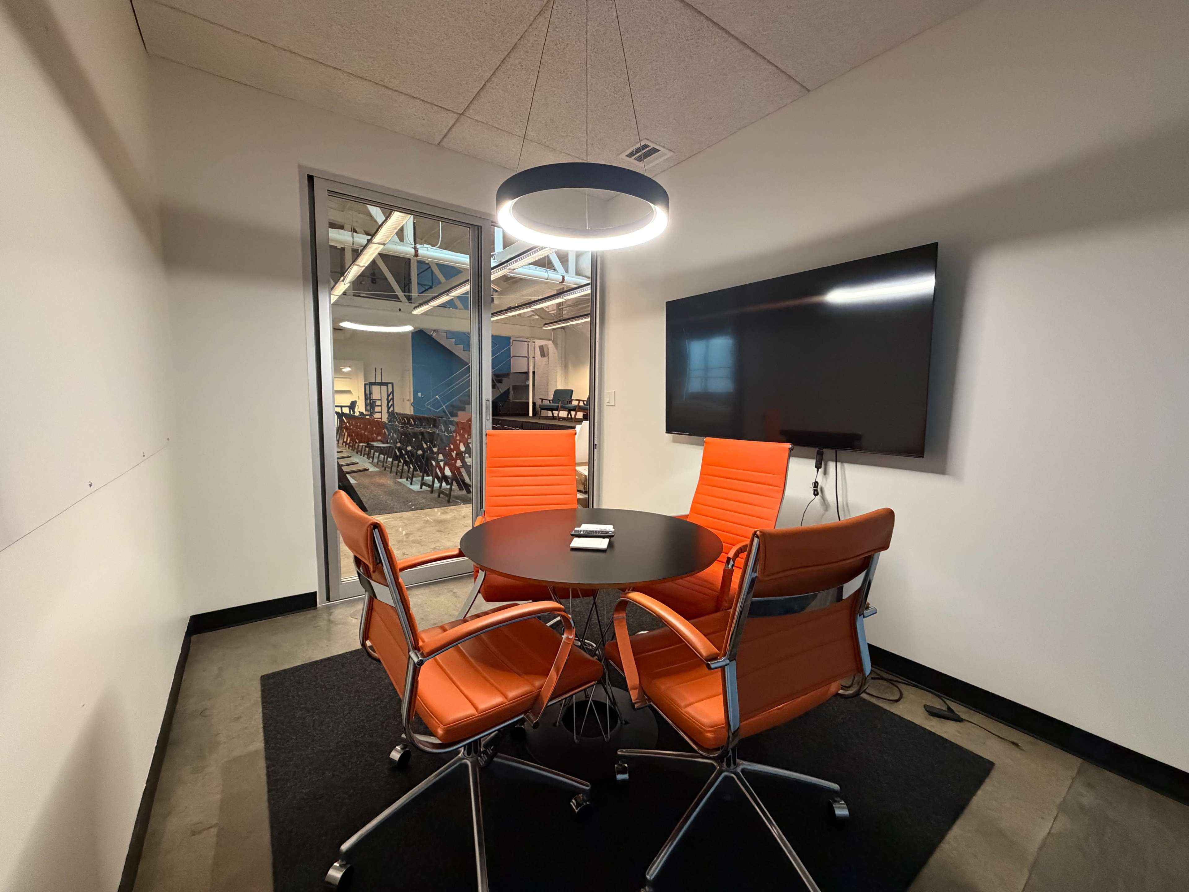 A small conference room features a round table surrounded by four orange chairs and a large screen on the wall.