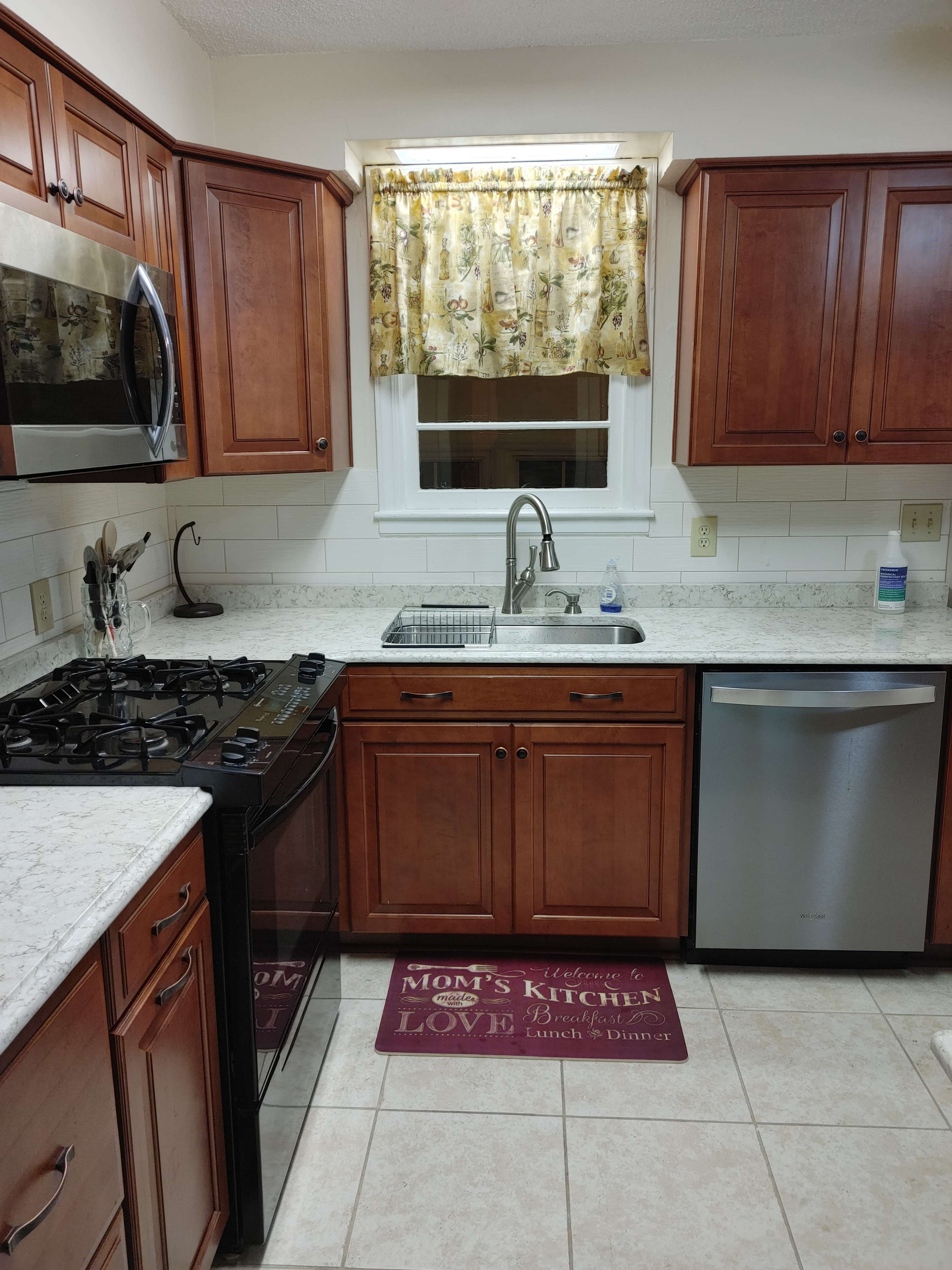 The image shows a kitchen with wooden cabinets, a sink beneath a window with a floral curtain, a stove, and a stainless steel dishwasher.