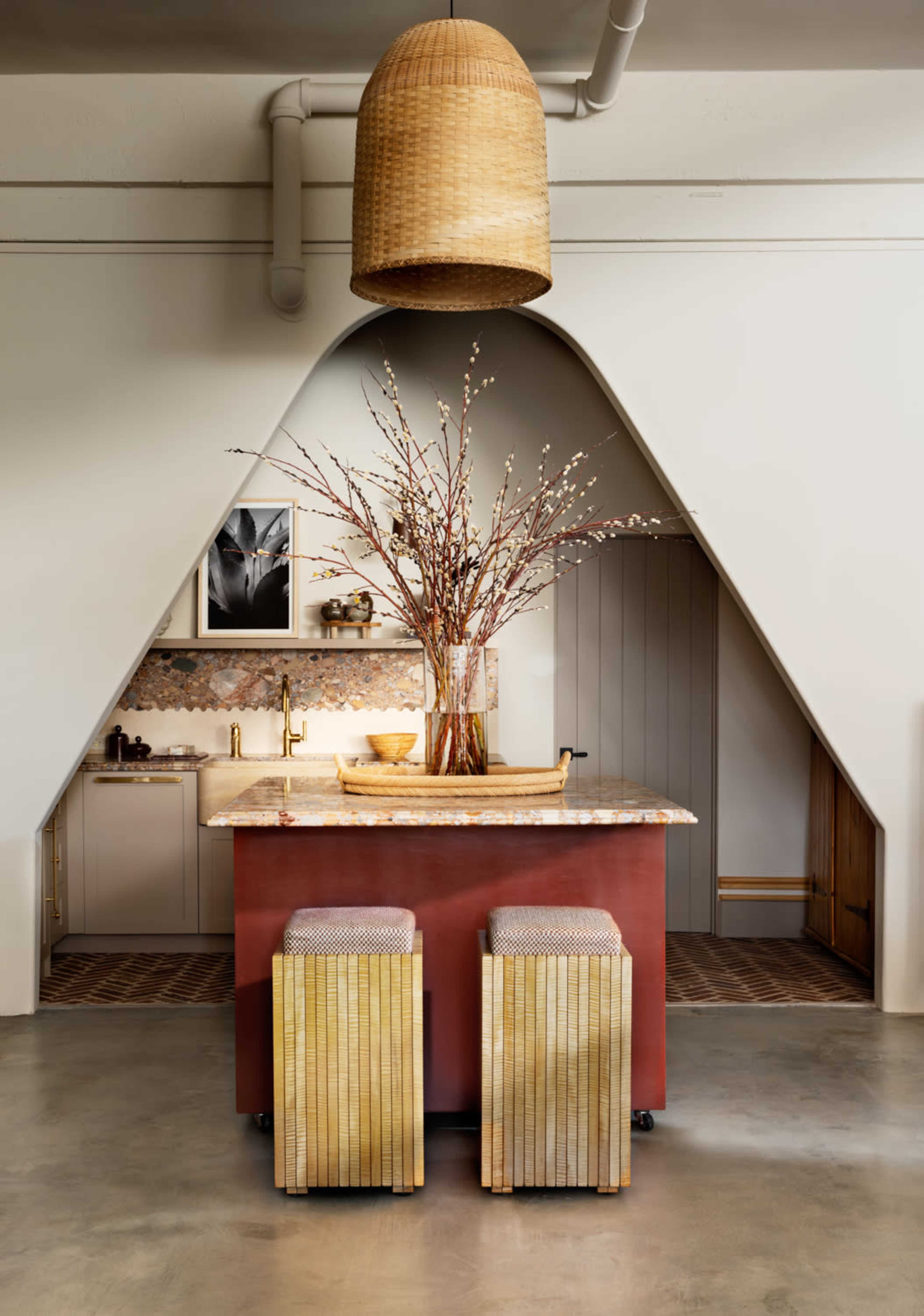 A modern kitchen features a central island with a patterned countertop, accompanied by two textured stools and a large woven pendant light overhead.