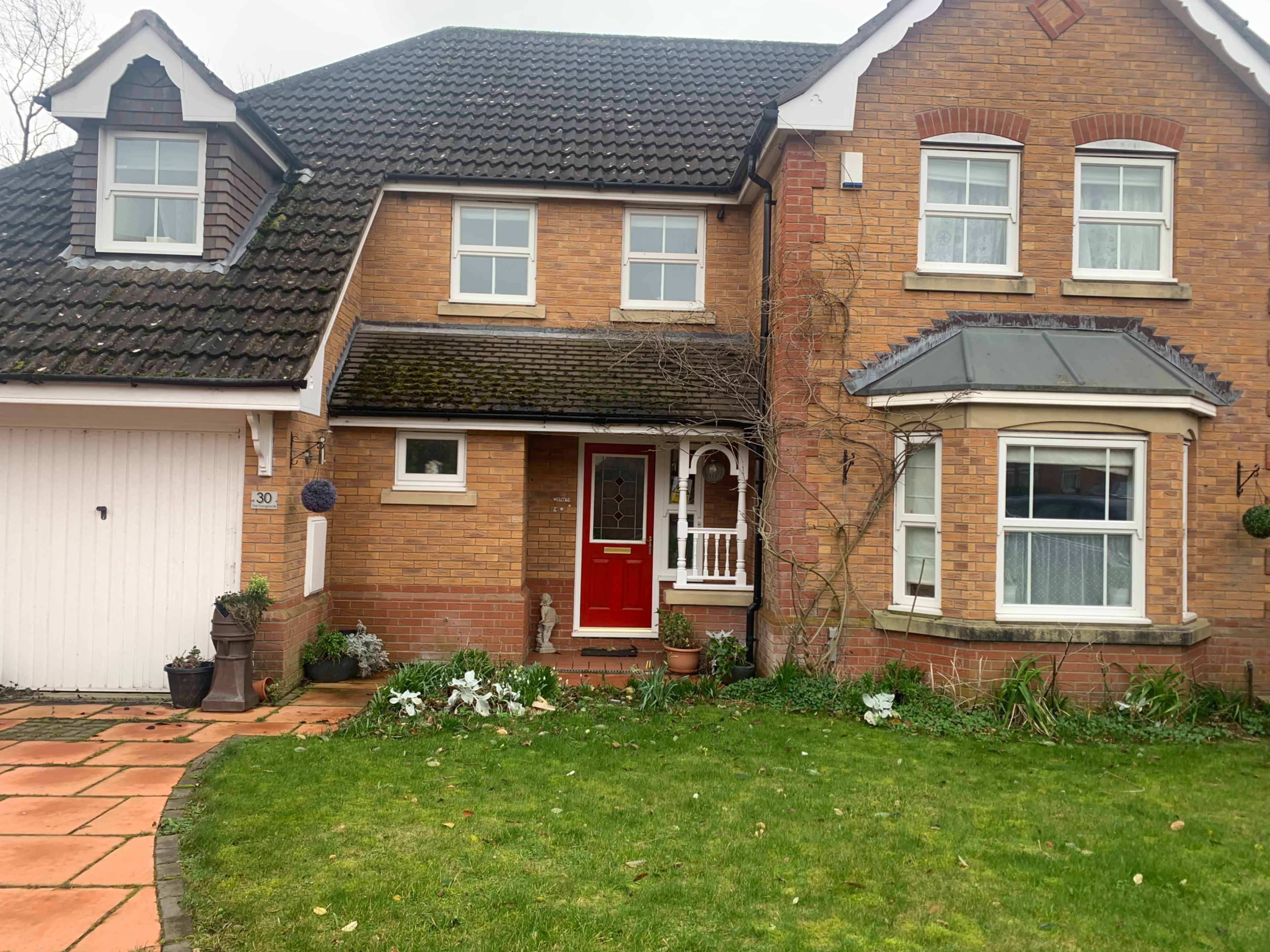 The image shows a brick-fronted residential house with a red door, surrounded by well-maintained gardens and a stone pathway.