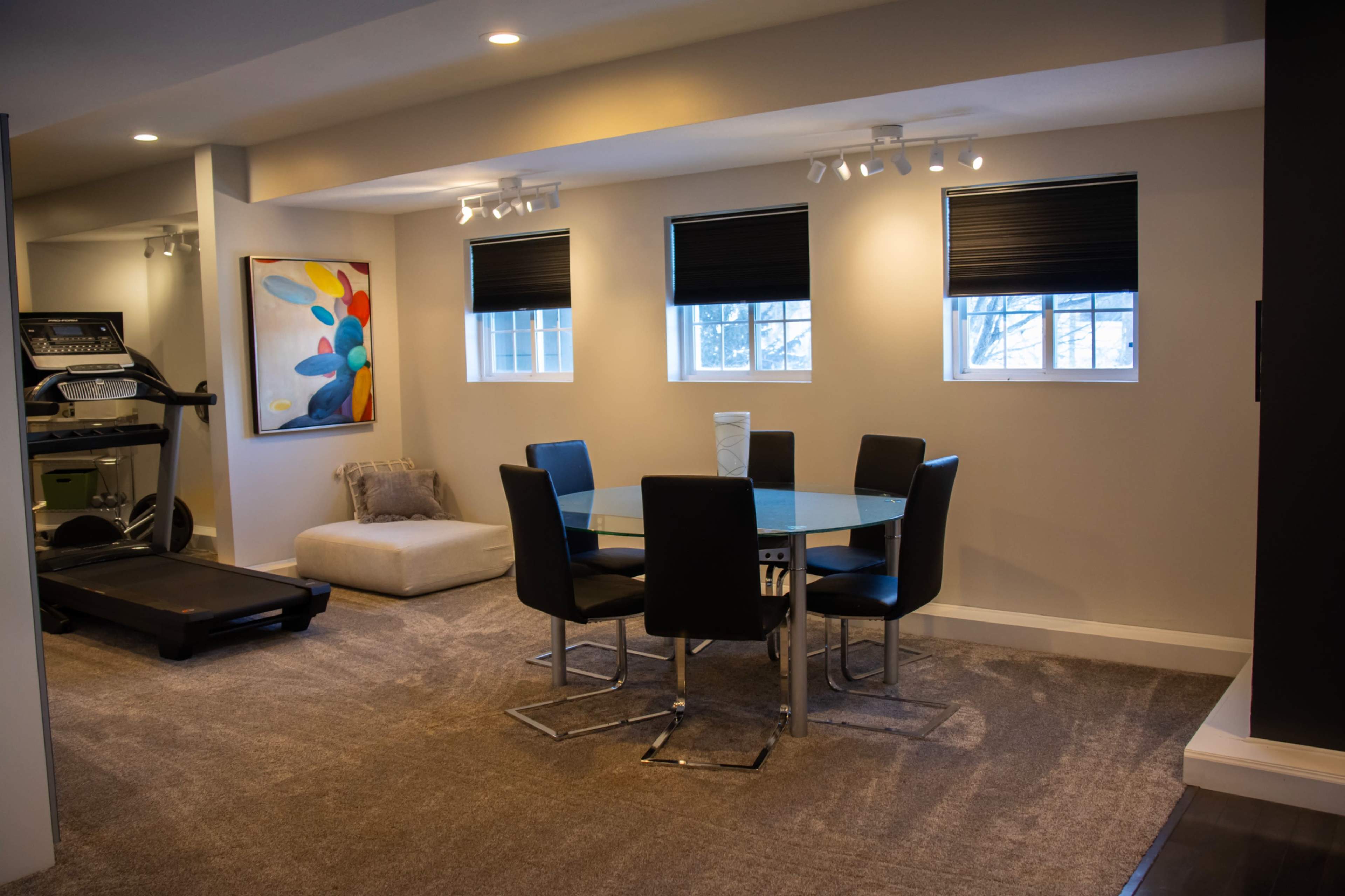 A modern dining area features a glass table surrounded by black chairs, with a treadmill and a sofa in the background.