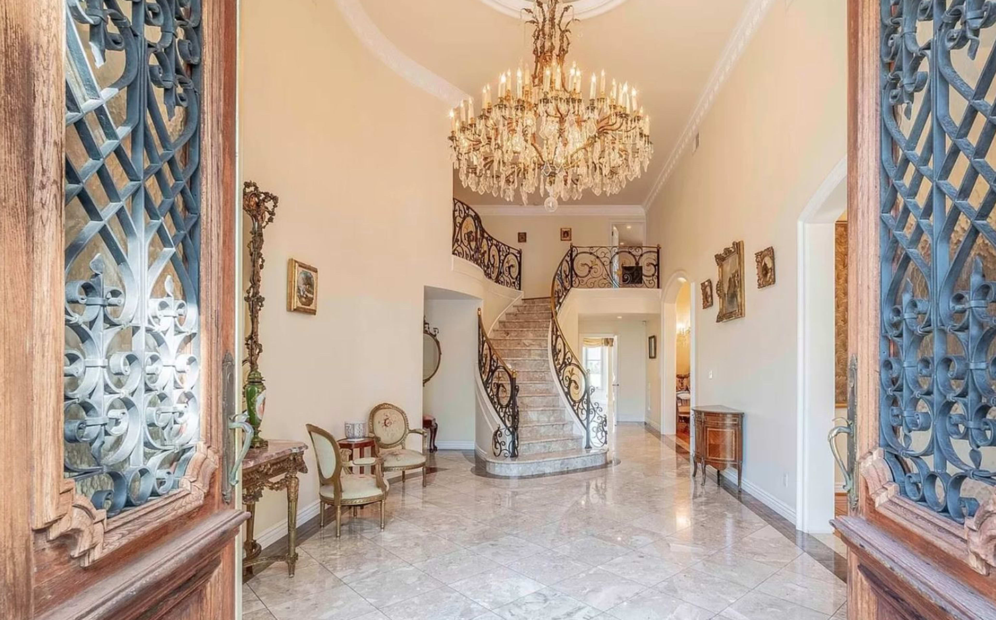 The image shows a grand foyer with a large chandelier, elegant staircase, and ornate ironwork doors.