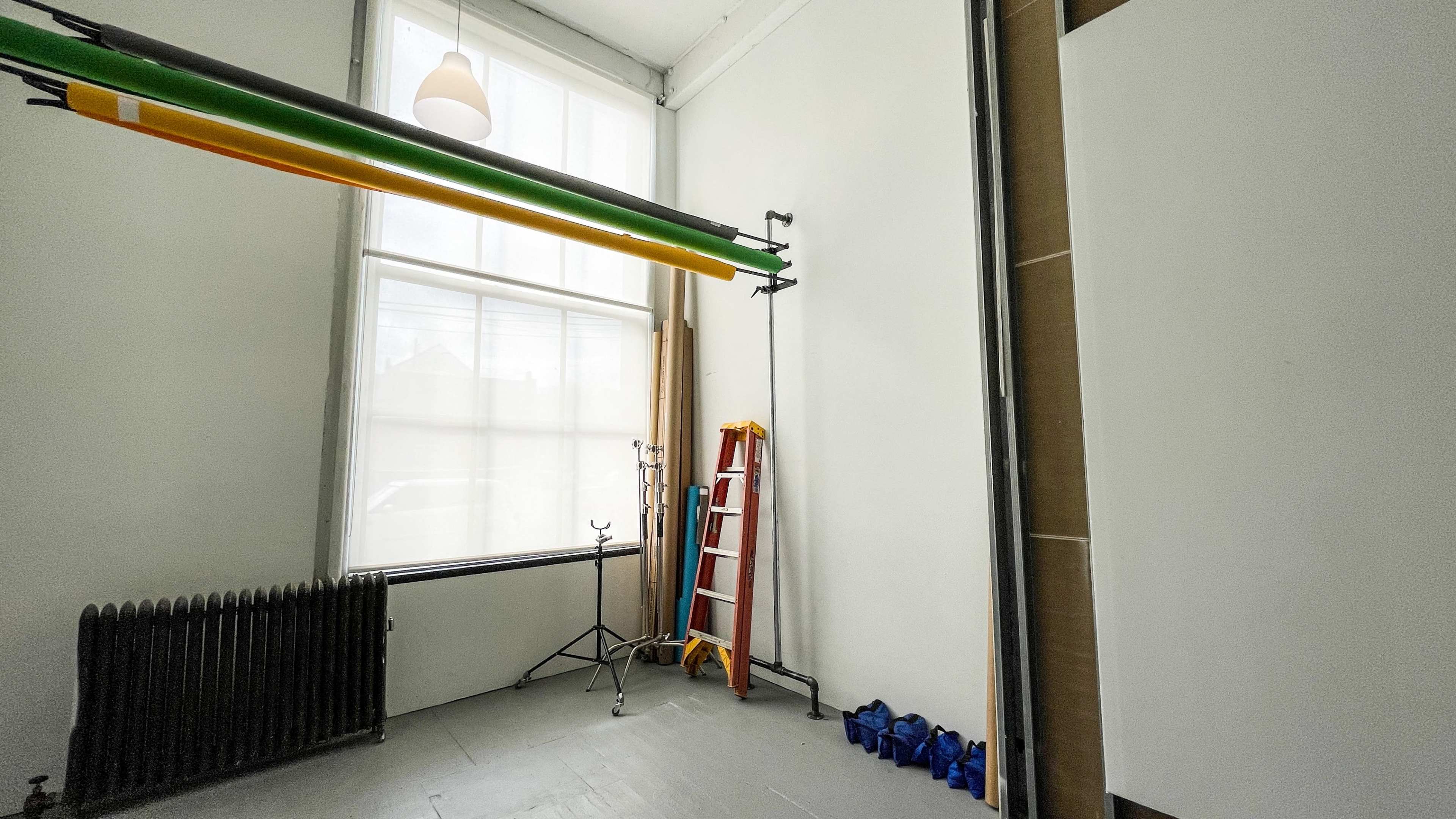 The image shows a brightly lit room with a ladder, backdrop poles with rolls of fabric, and equipment organized along the walls.