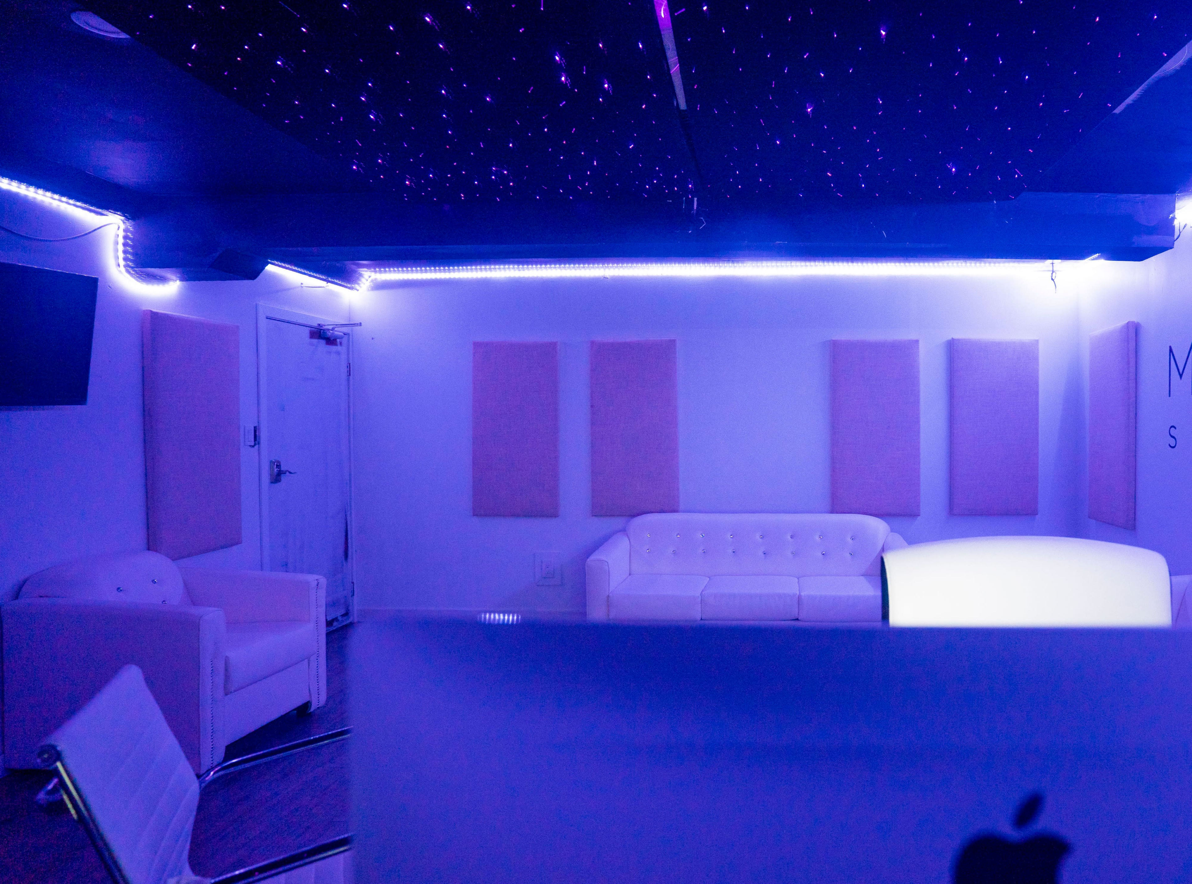 A modern, dimly lit studio space with a white couch, soundproof panels on the walls, and ambient purple lighting.