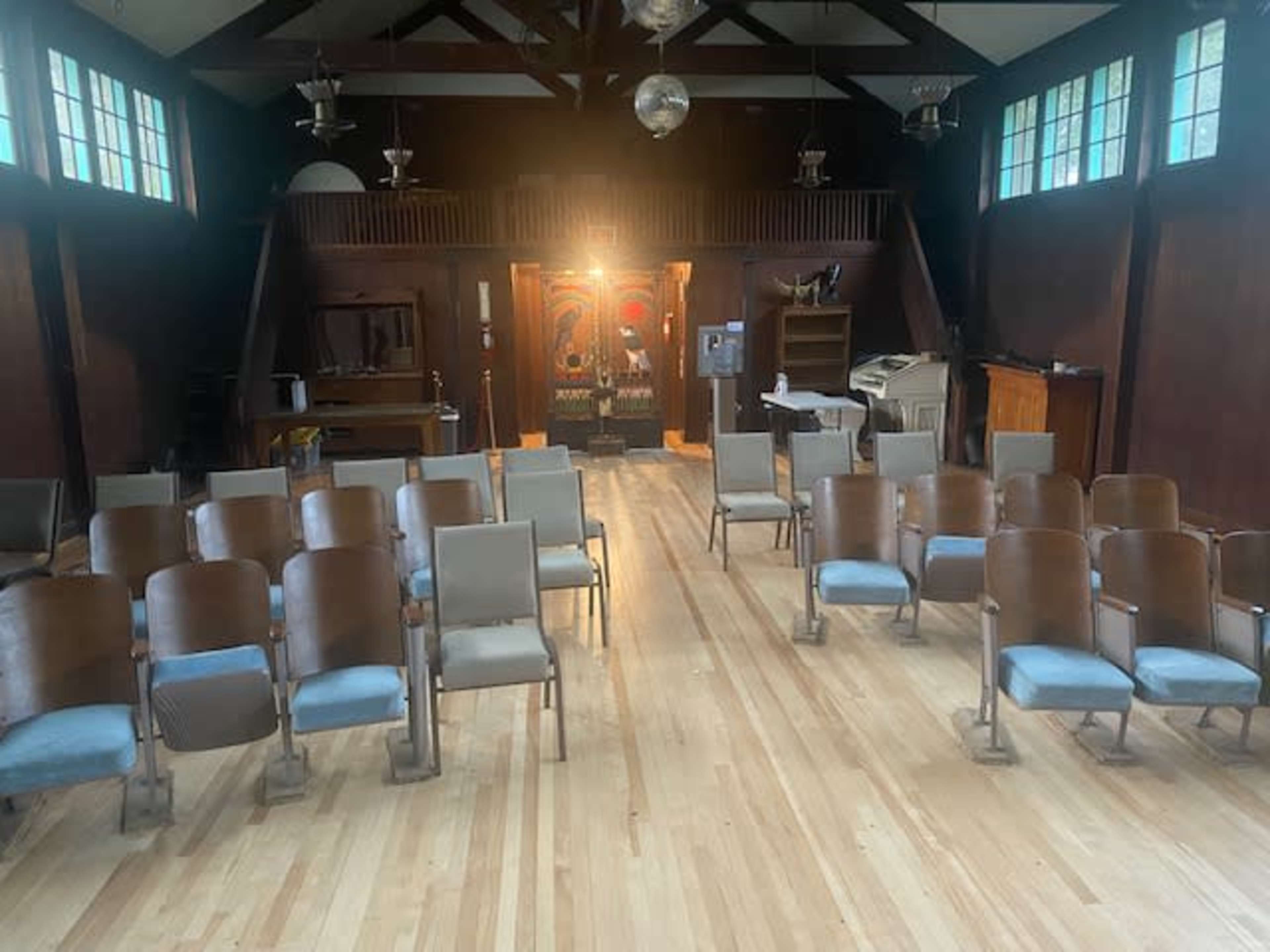 A wooden-floored room with rows of metal-framed chairs facing a stage and a brightly lit background.