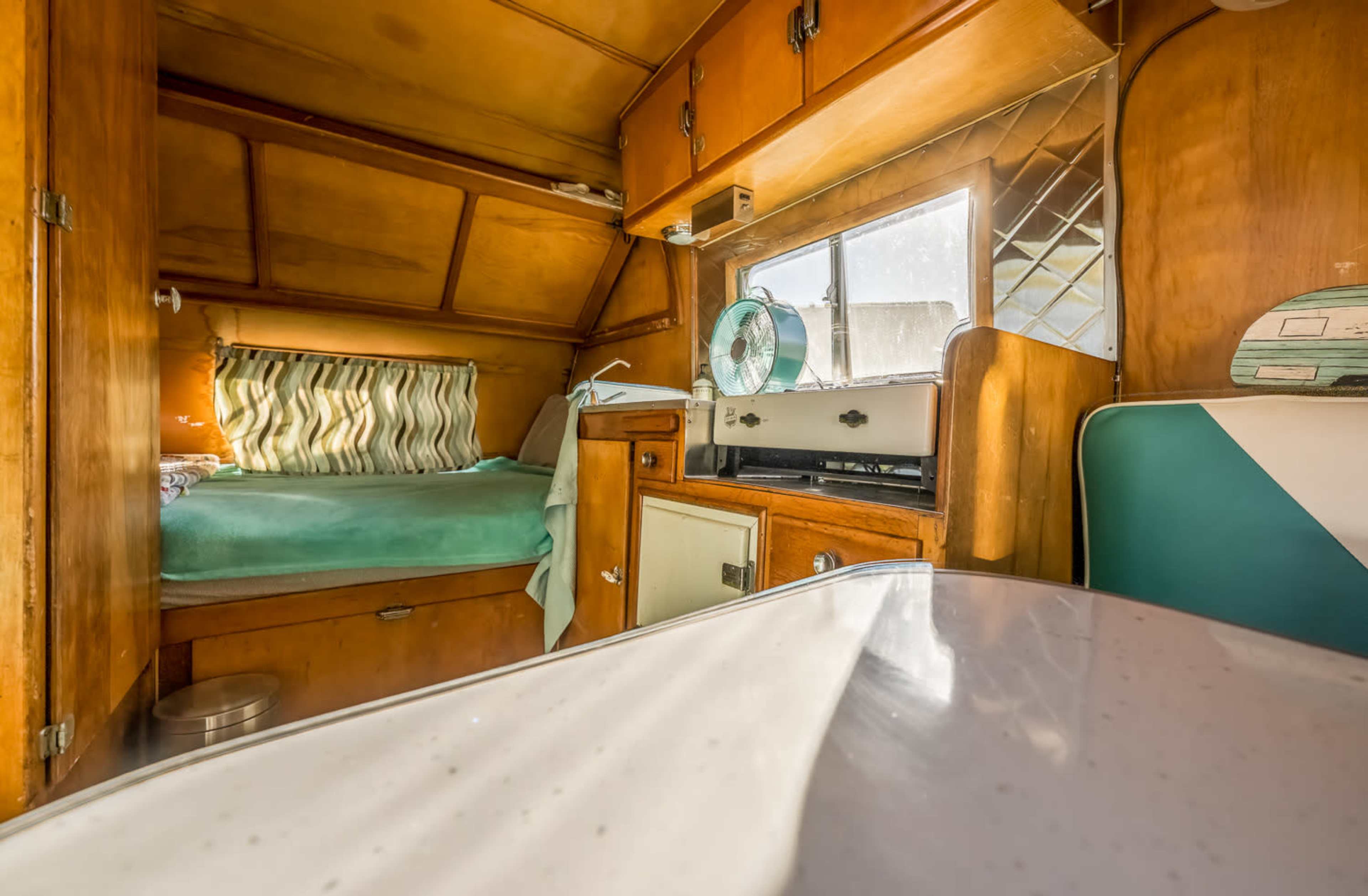 The interior of a vintage camper with wooden paneling, a small bed, and a kitchenette area featuring a window and a green fan.
