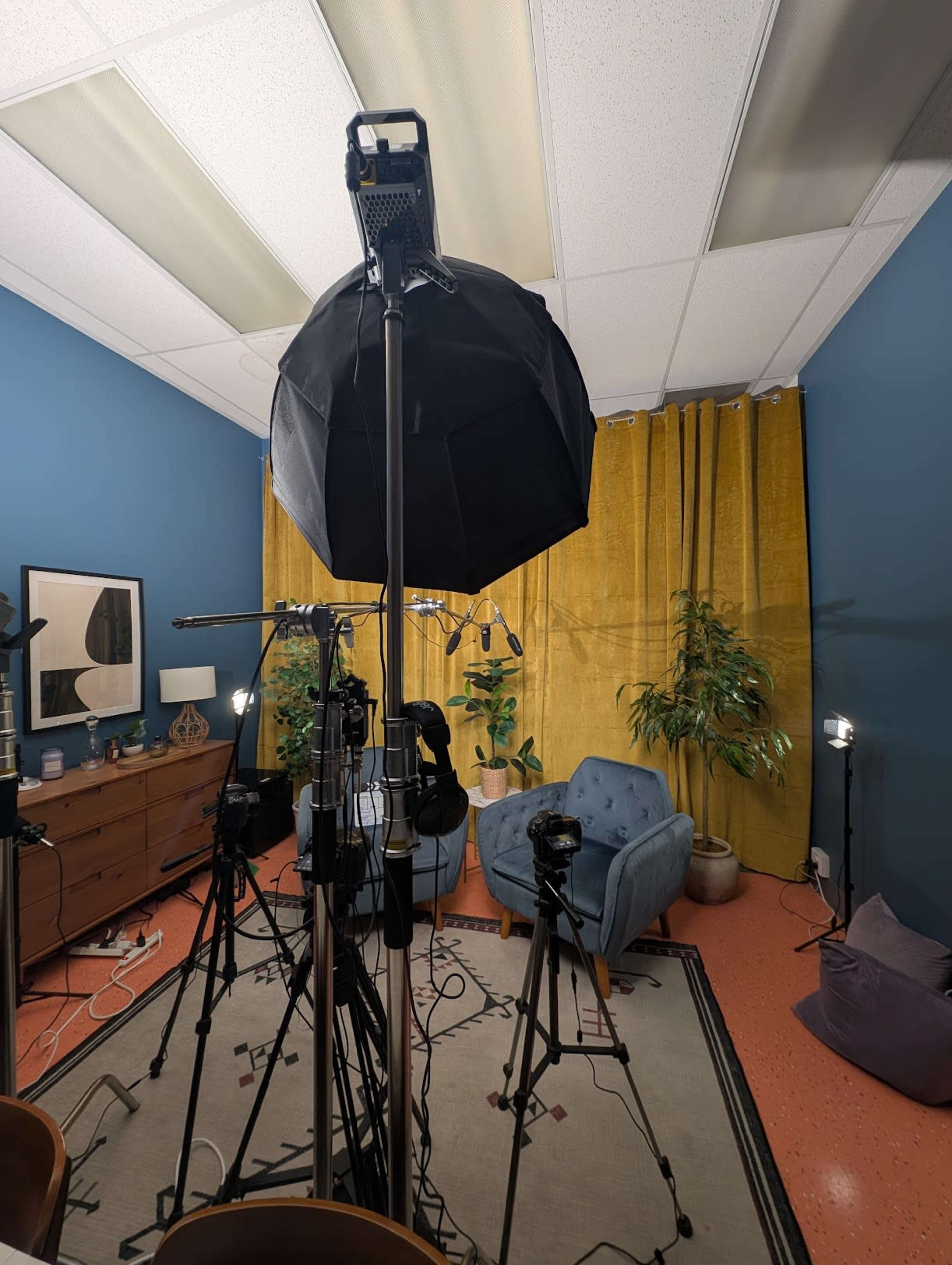 The image shows a studio setup with multiple cameras and lighting equipment focused on a blue armchair surrounded by plants and yellow curtains.