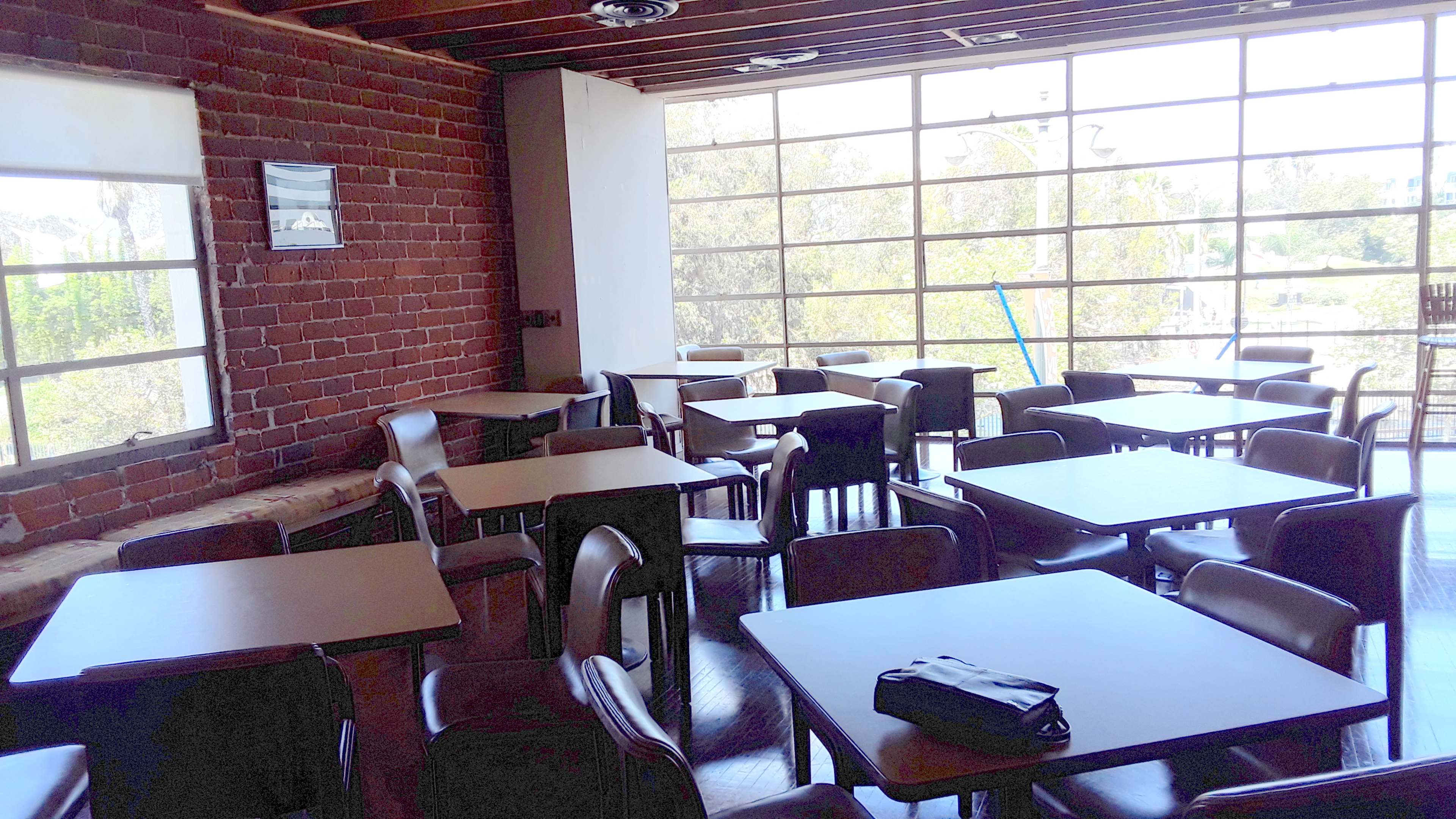 A dining area featuring several empty tables and chairs arranged in a spacious room with large windows and a brick wall.