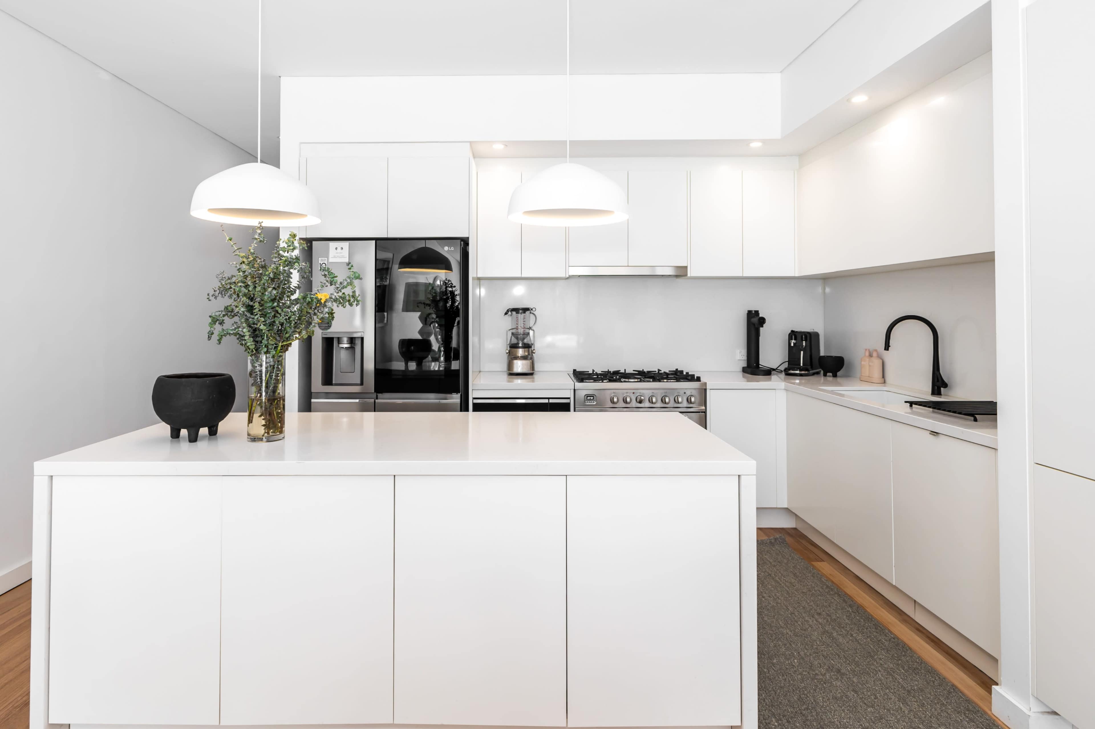 The image features a modern kitchen with white cabinetry, stainless steel appliances, and a central island with a vase of greenery.