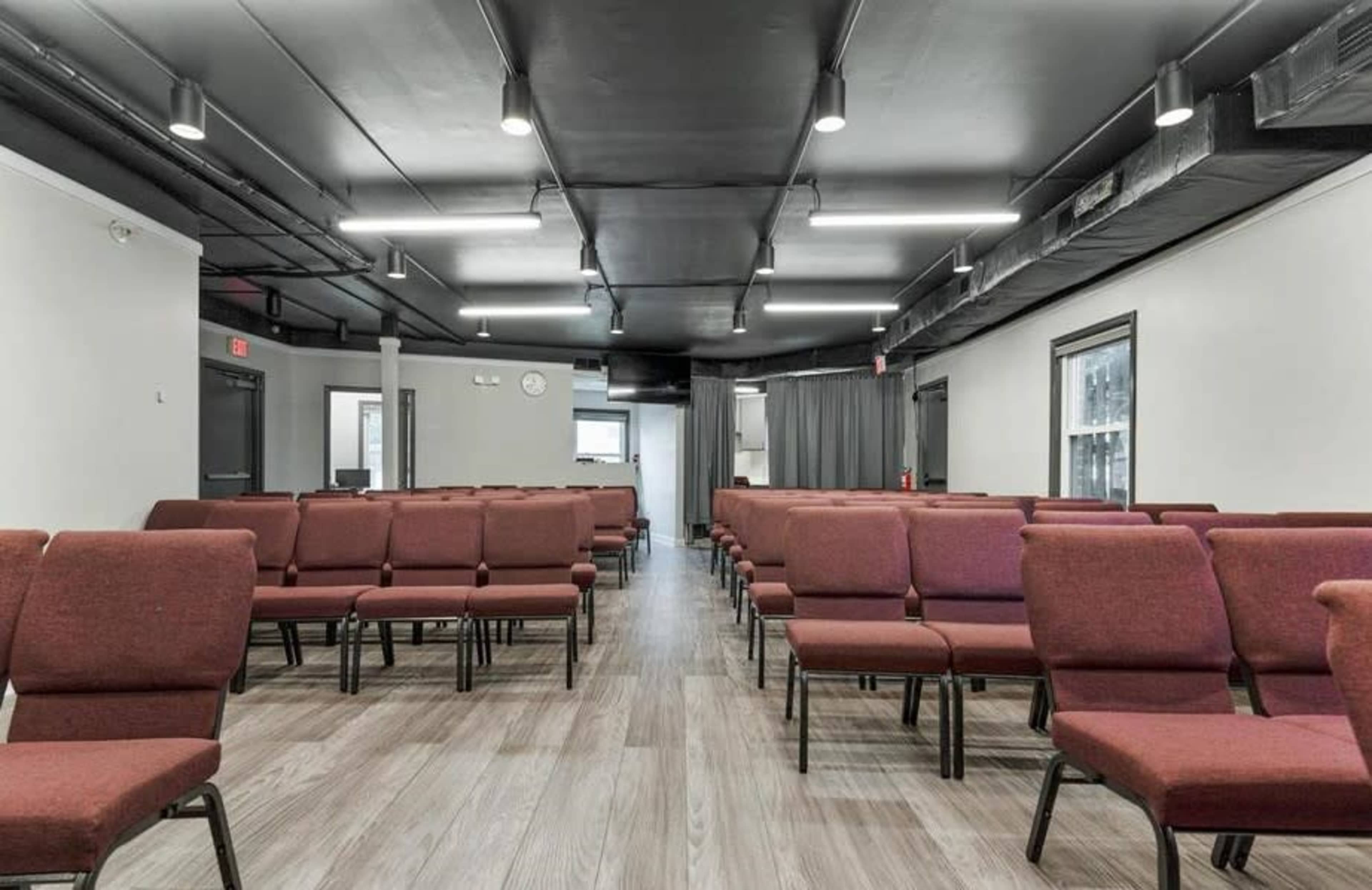 A spacious room with rows of red chairs arranged facing the front, illuminated by ceiling lights and featuring a gray floor.