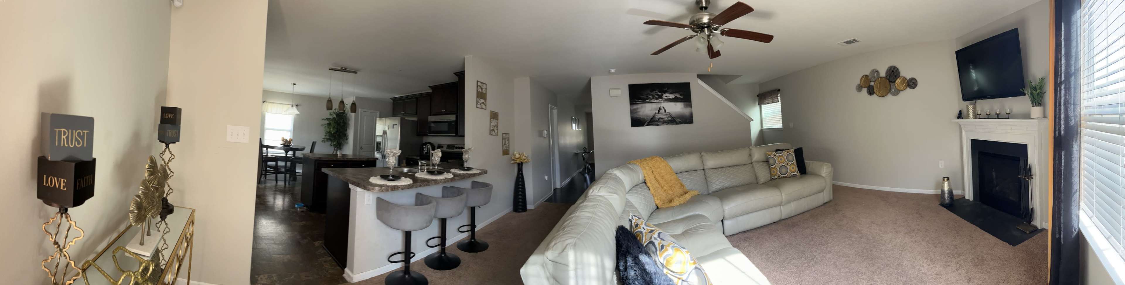 A modern living space features a spacious open-concept layout with a gray sectional sofa, a kitchen bar with stools, and a dining area in the background.