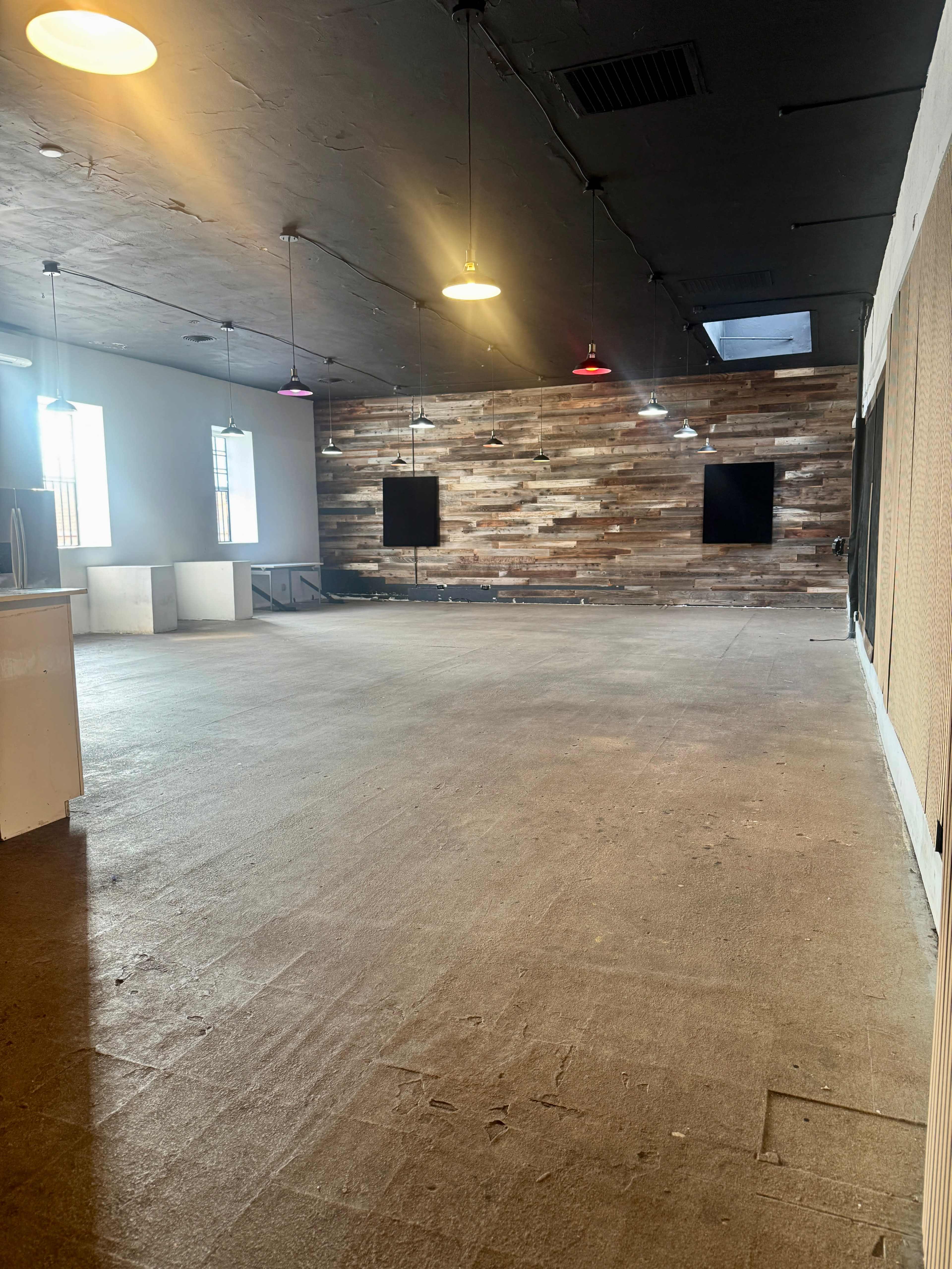 The image shows an empty interior space with a wooden accent wall, overhead pendant lighting, and a concrete floor.