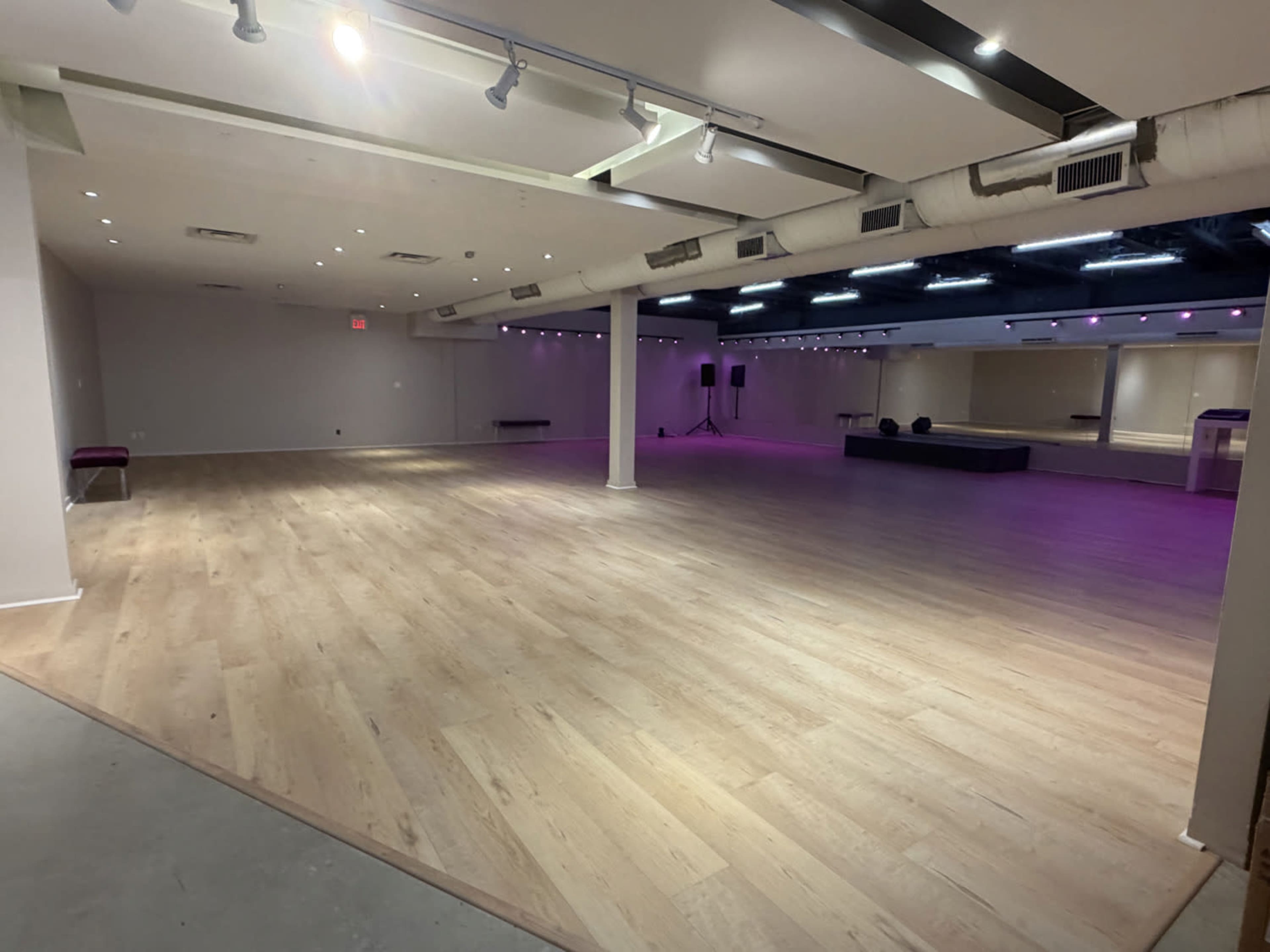The image shows a spacious, well-lit studio with wooden flooring and purple lighting along the ceiling, featuring a mirrored wall and a small stage area.