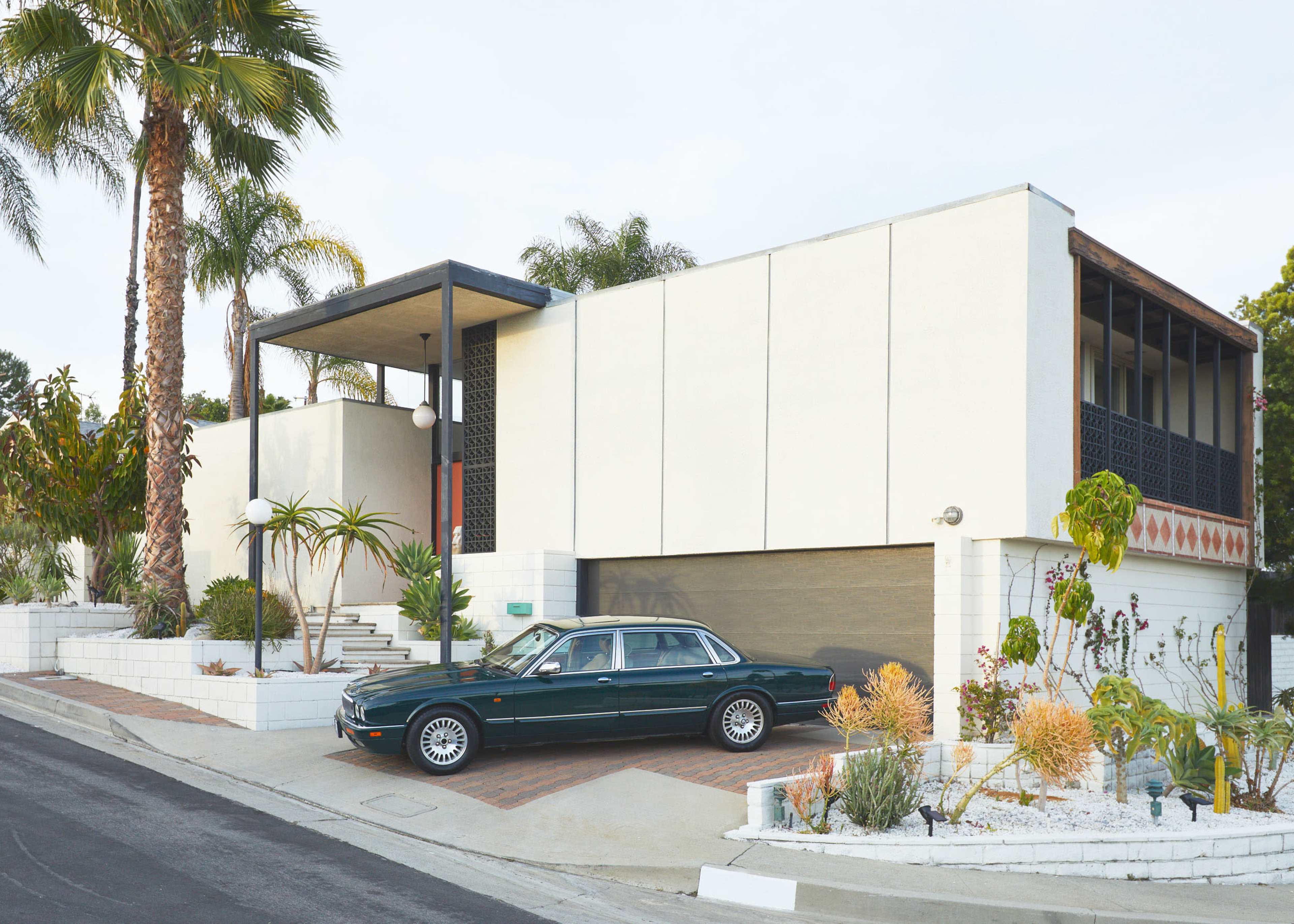A modern two-story house with a green car parked in front, featuring palm trees and various desert plants in the landscaping.