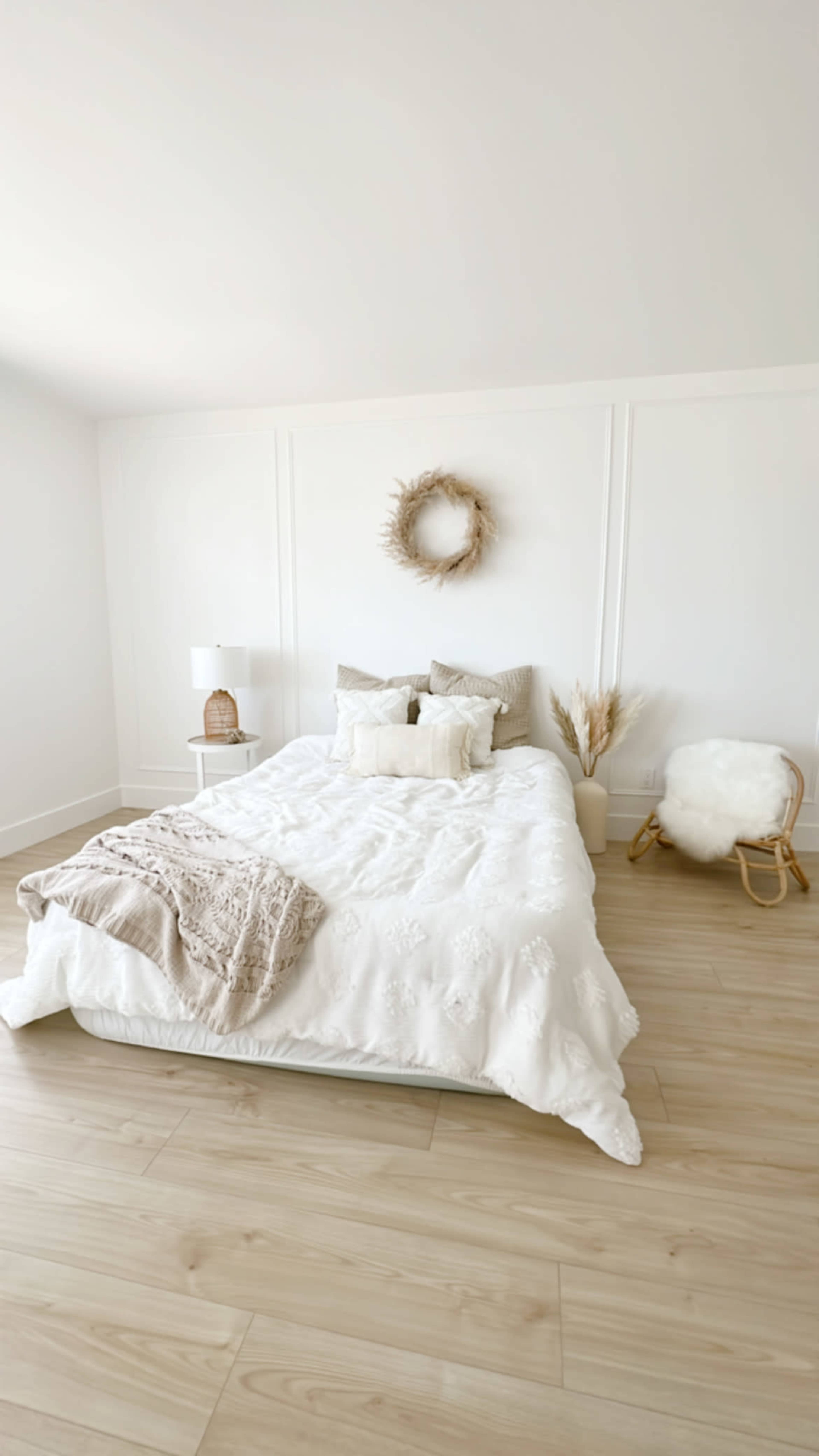 A bright, minimalist bedroom with a large bed covered in white bedding, a decorative wreath on the wall, and a cozy armchair in the corner.