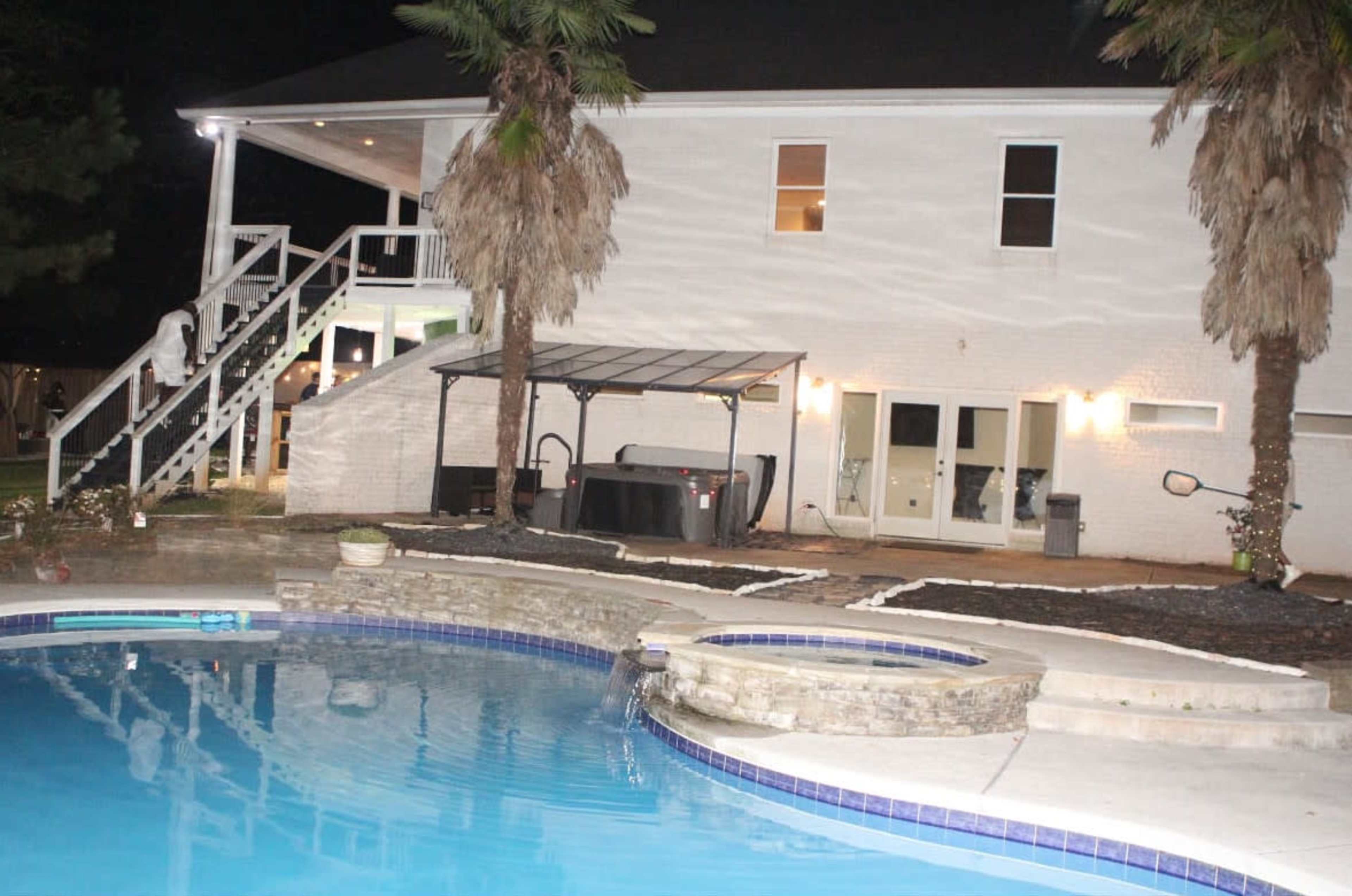 The image shows a lit backyard at night featuring a swimming pool, a large house with patio stairs, and a hot tub under a gazebo.