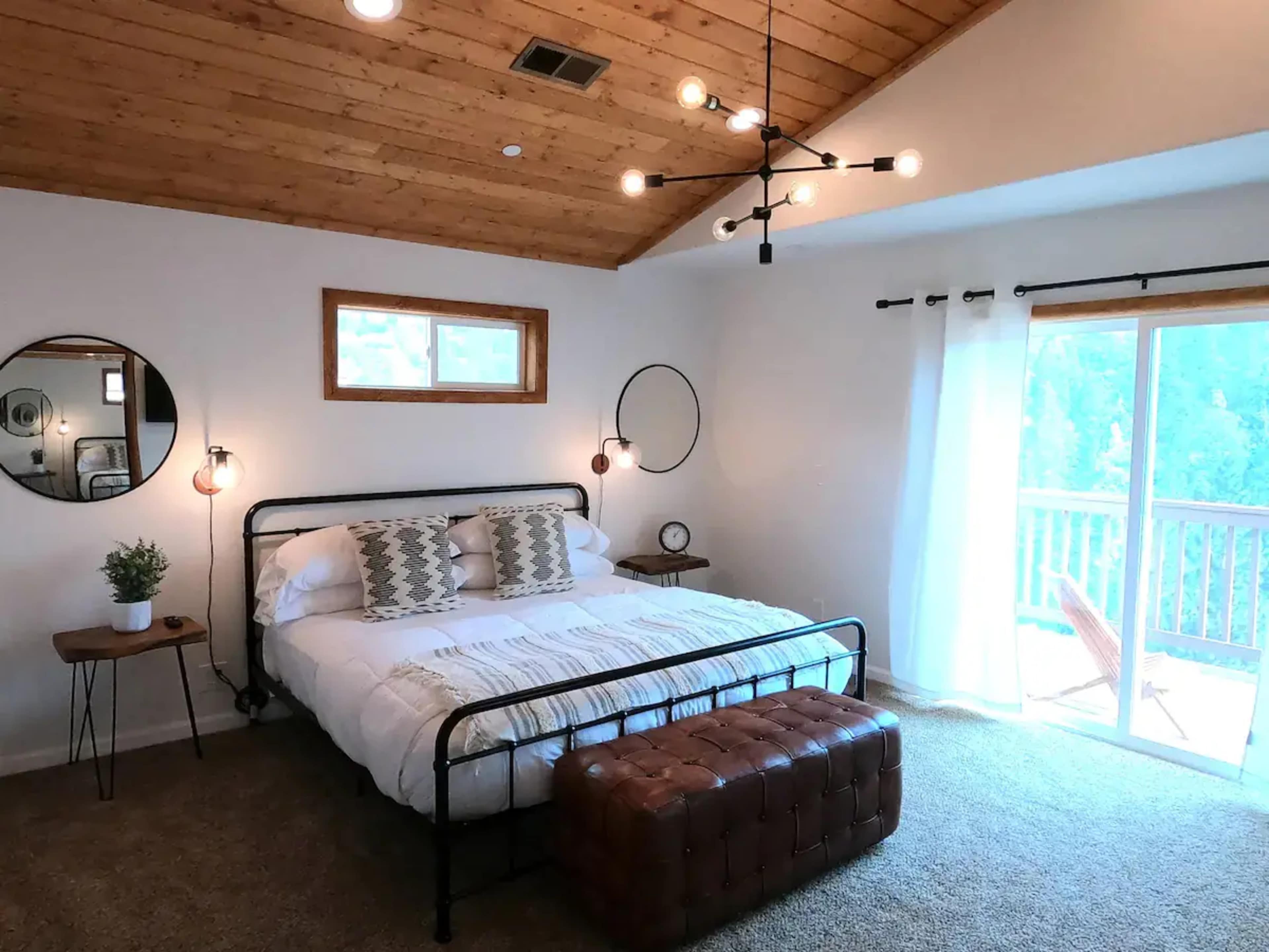 The image shows a spacious bedroom with a large bed, a wooden ceiling, a round mirror, and sliding glass doors leading to a balcony.