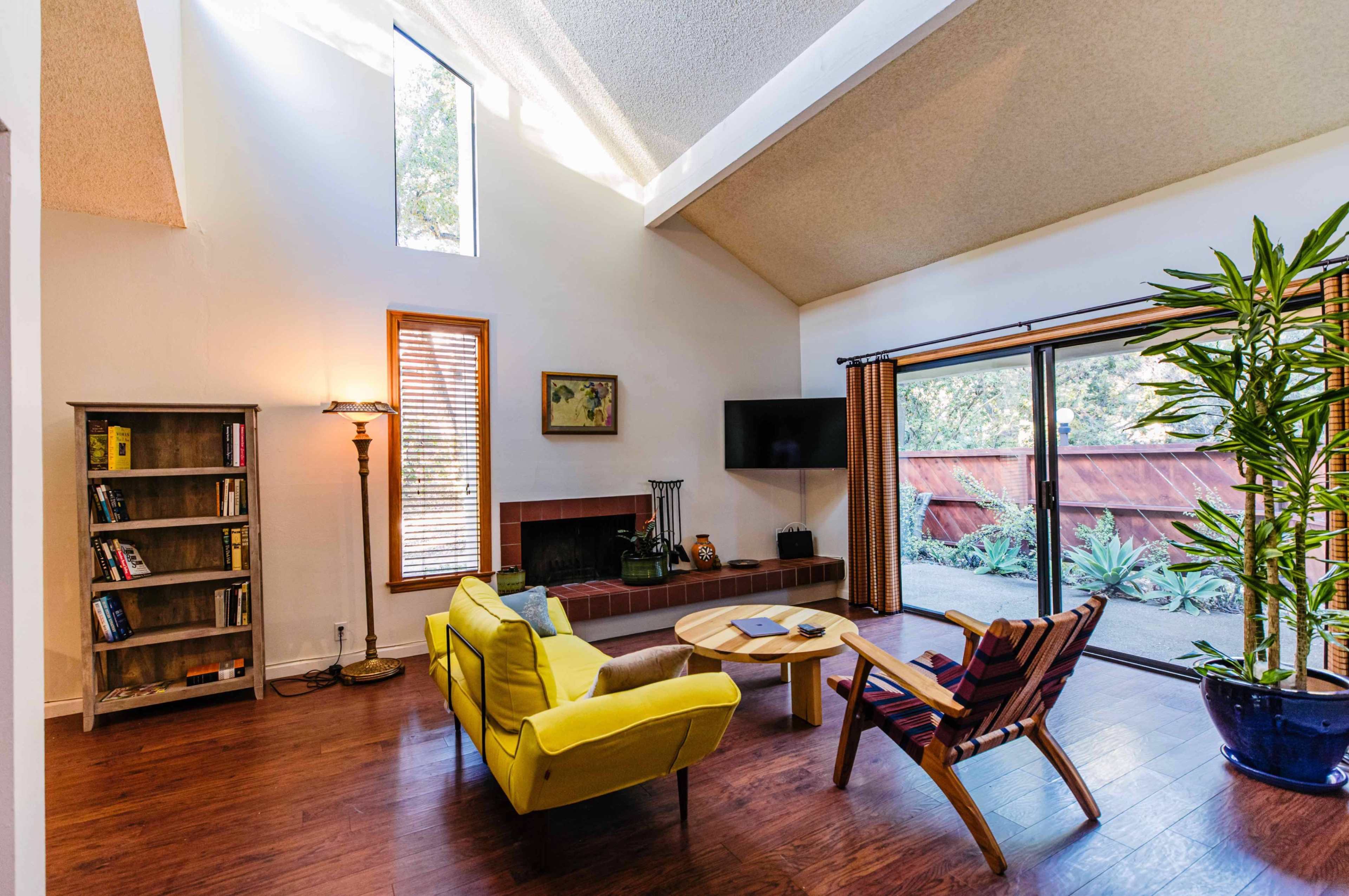 A bright living room features yellow sofas, a wooden coffee table, a bookshelf, and large sliding glass doors leading to an outdoor space.