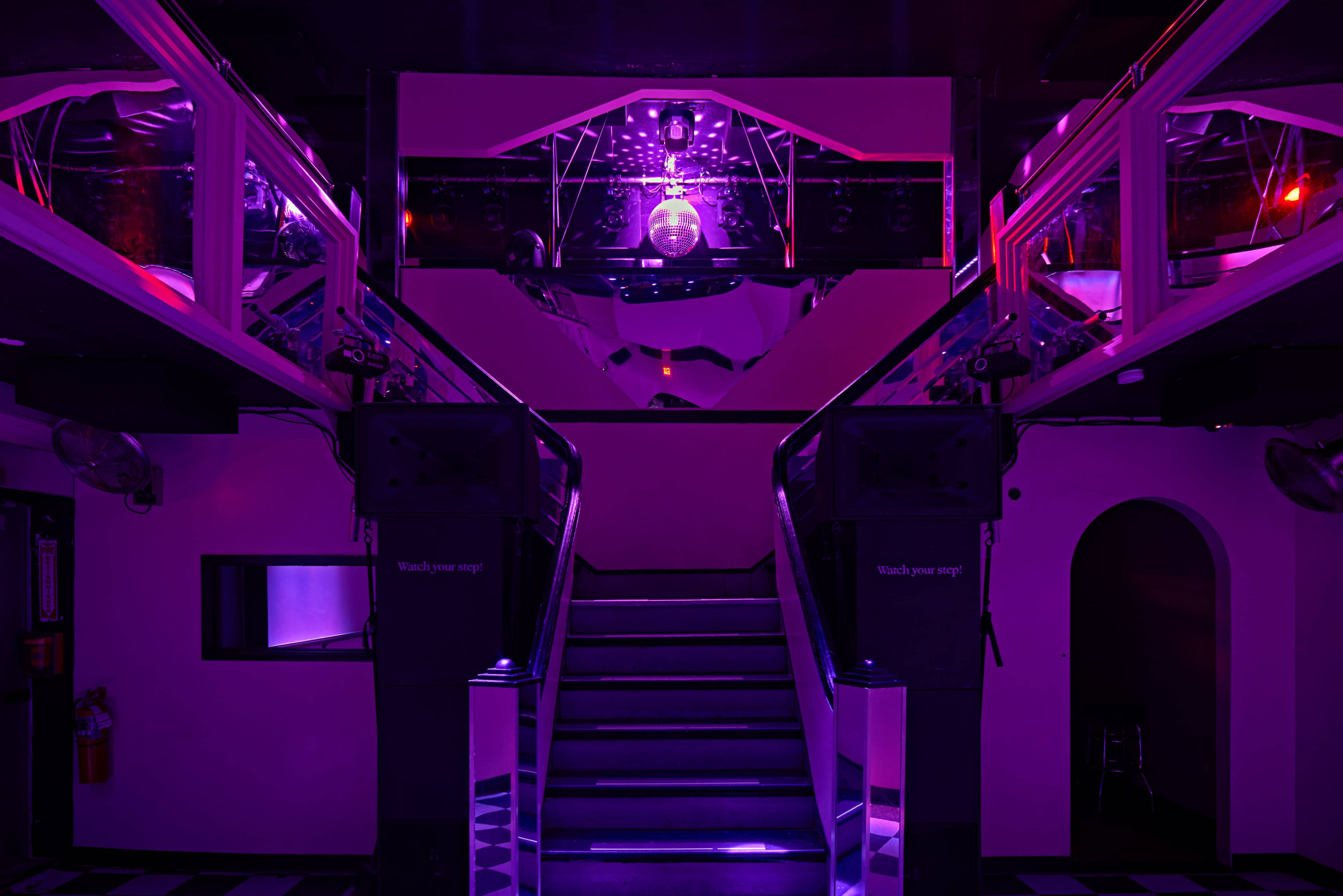 A dimly lit nightclub interior with a staircase leading to a disco ball and illuminated DJ booth, surrounded by speakers and vibrant purple lighting.