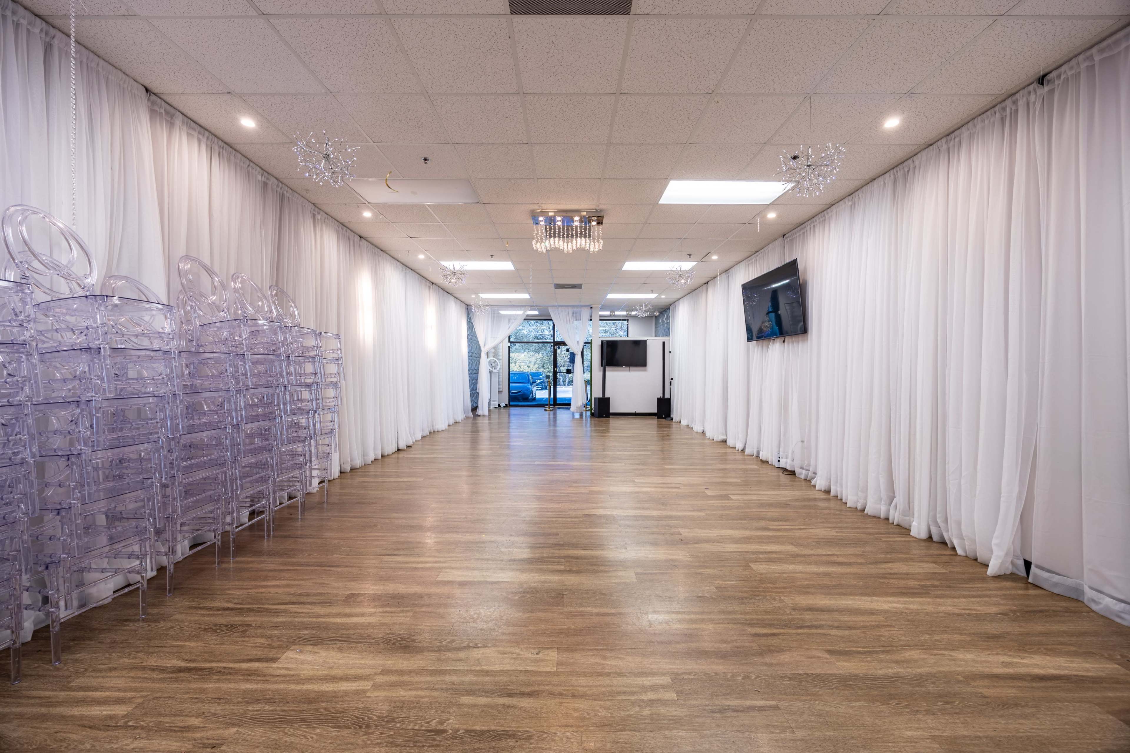 The image shows a spacious interior room with a wooden floor, lined with white curtain drapes, and featuring a stack of transparent chairs on one side and a chandelier on the ceiling.
