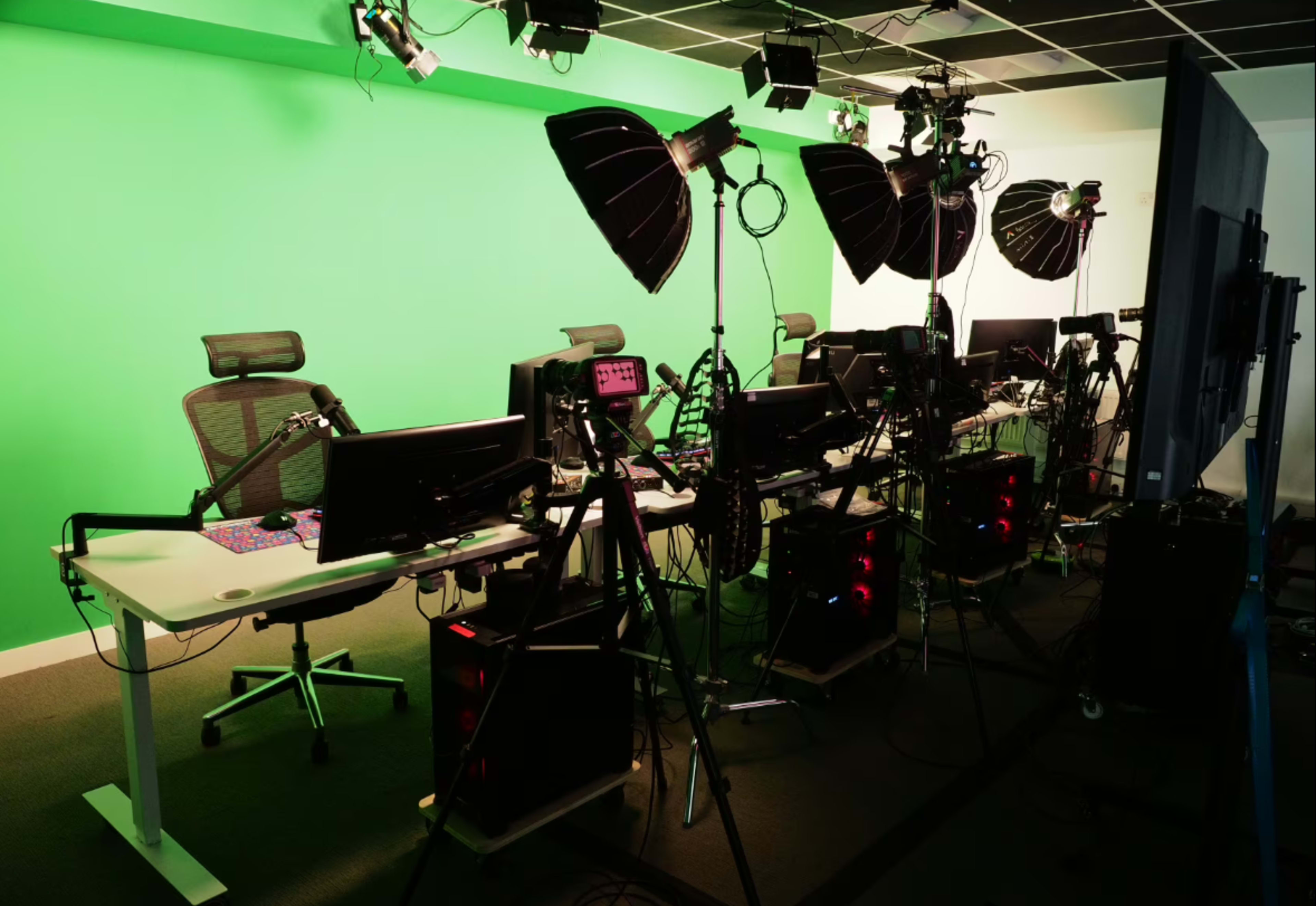 The image shows a studio setup with multiple workstations, green screens, lighting equipment, and computers arranged in a row.