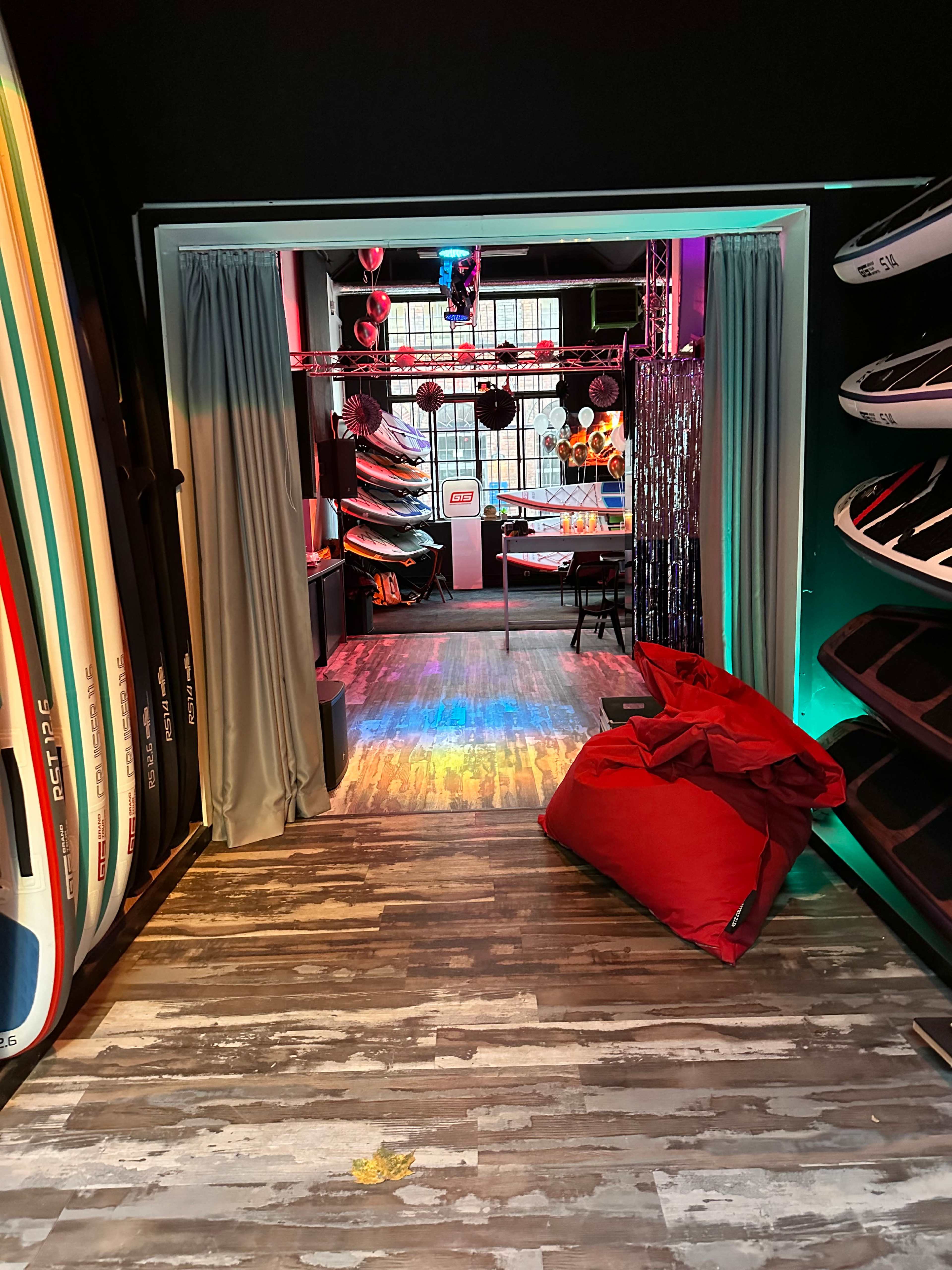 The image shows a dimly lit recreational space with colorful lighting, featuring surfboards hanging on the walls, and a lounge area with red bean bags and a table.