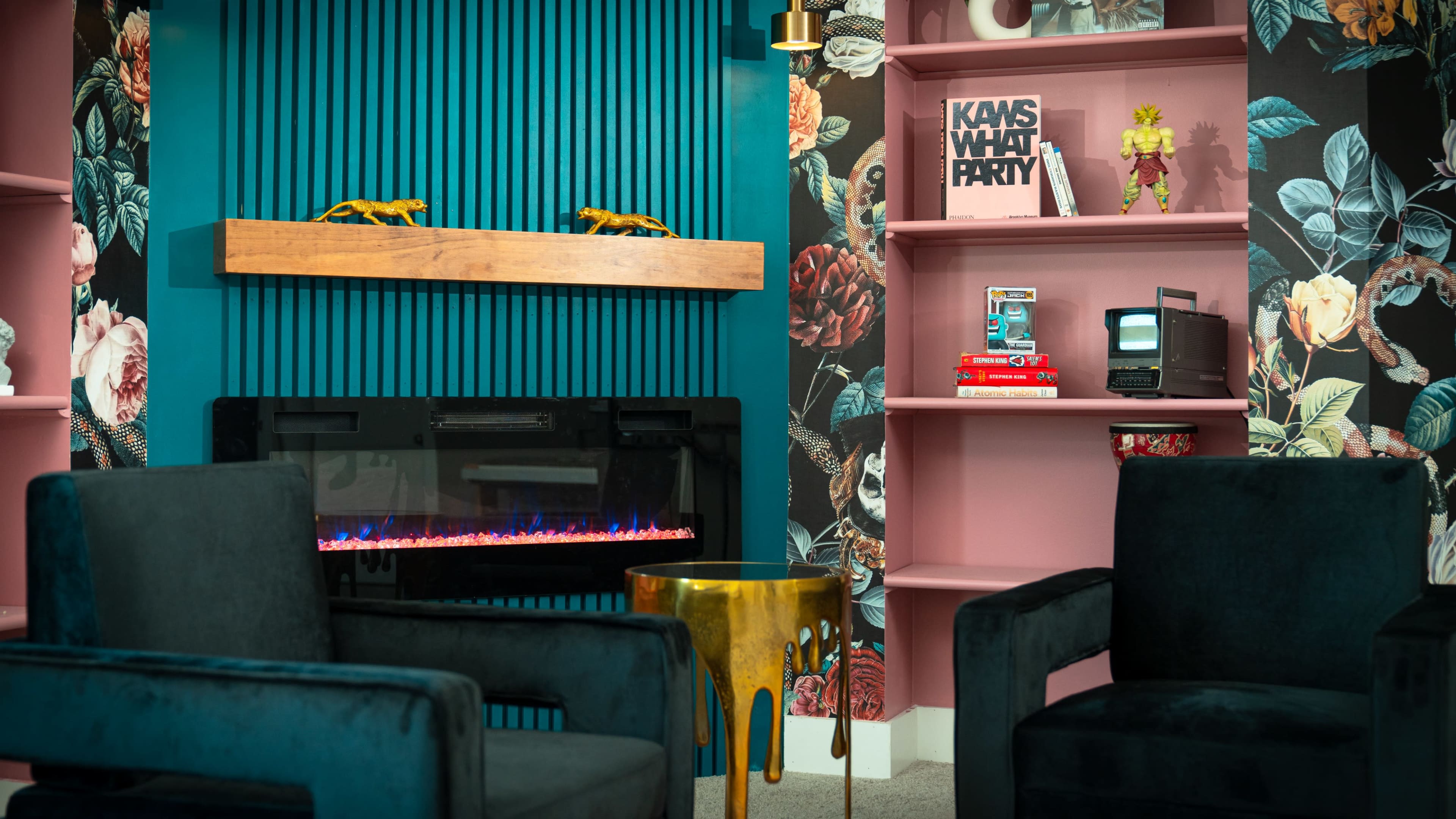 The image shows a modern living room with two velvet chairs facing a wall-mounted electric fireplace and a bookshelf featuring decorative items and a vintage television.