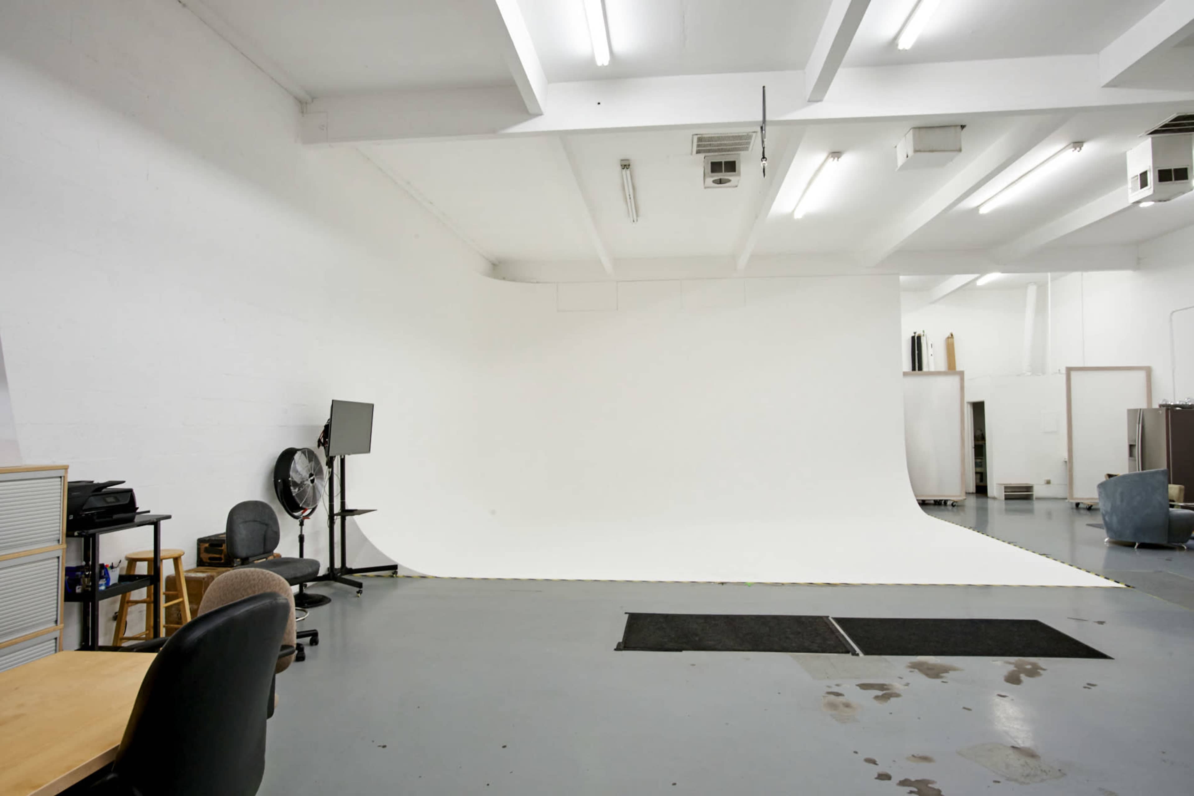A spacious, empty studio with a white backdrop, gray floor, and minimal furnishings including chairs and a desk.