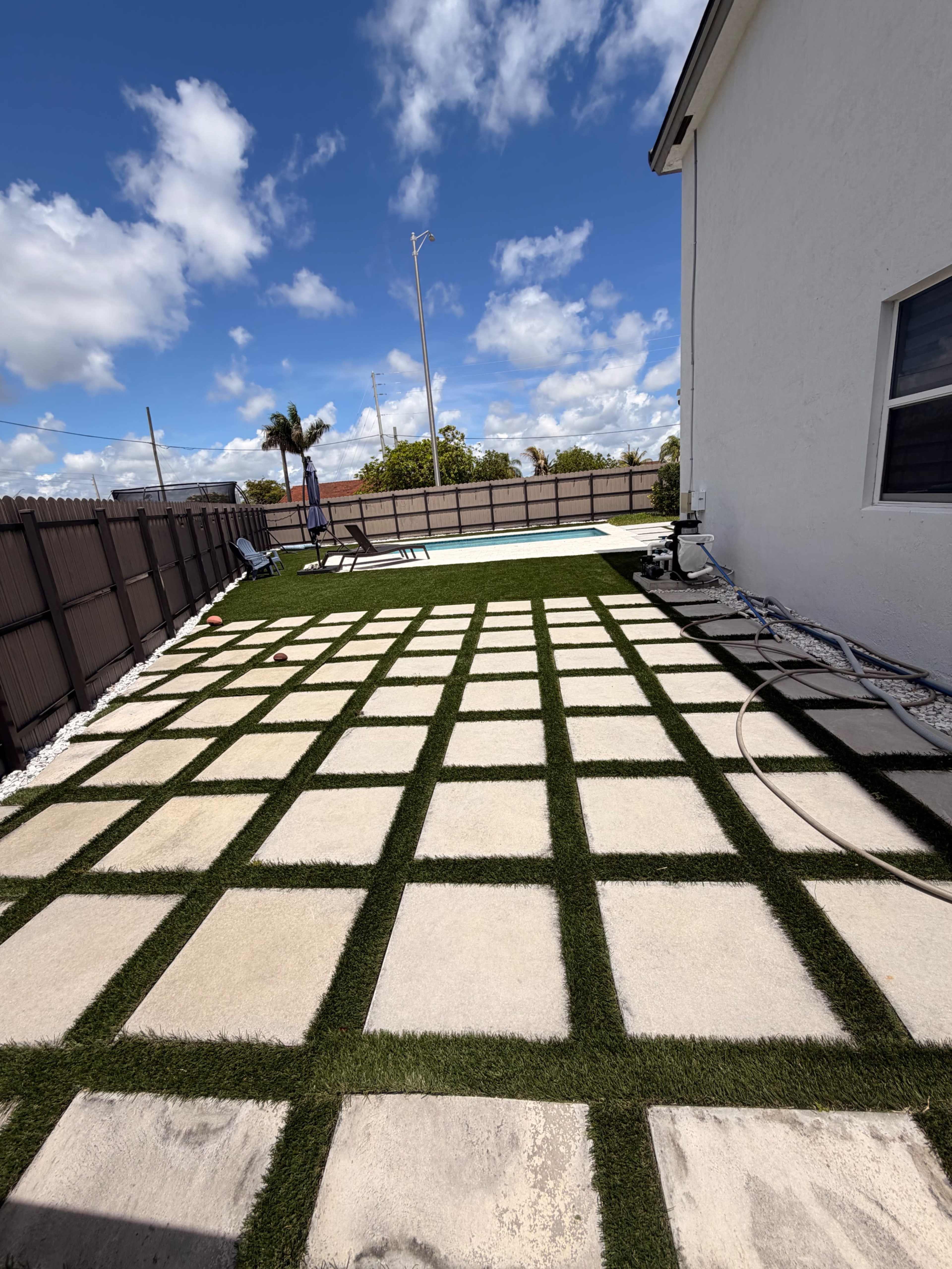 The image shows a backyard patio with large stone pavers arranged in a grid pattern over green artificial grass, adjacent to a swimming pool.