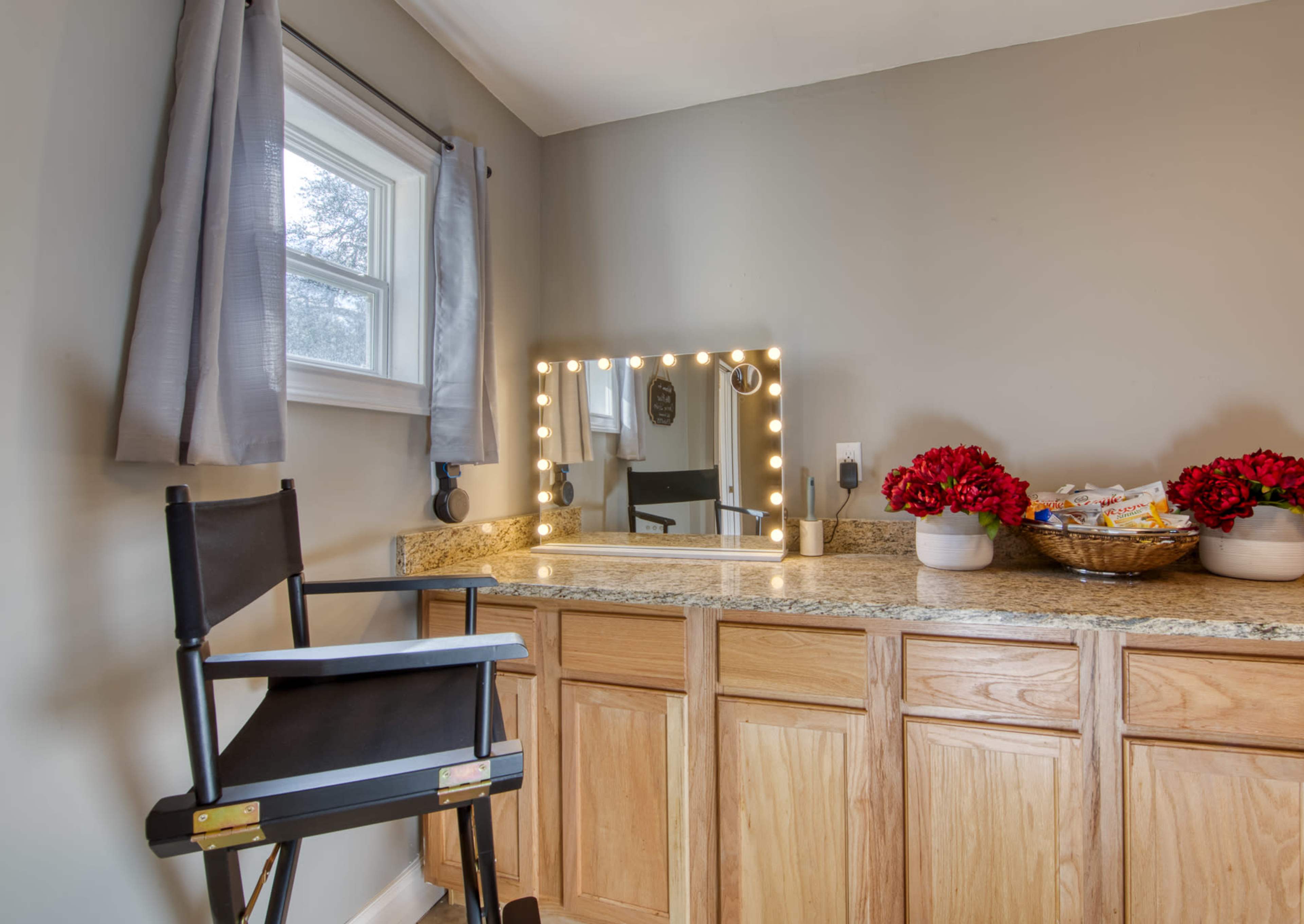 A makeup station is set up with a lighted mirror, granite countertop, and a chair beside a window, adorned with flowers in a basket.
