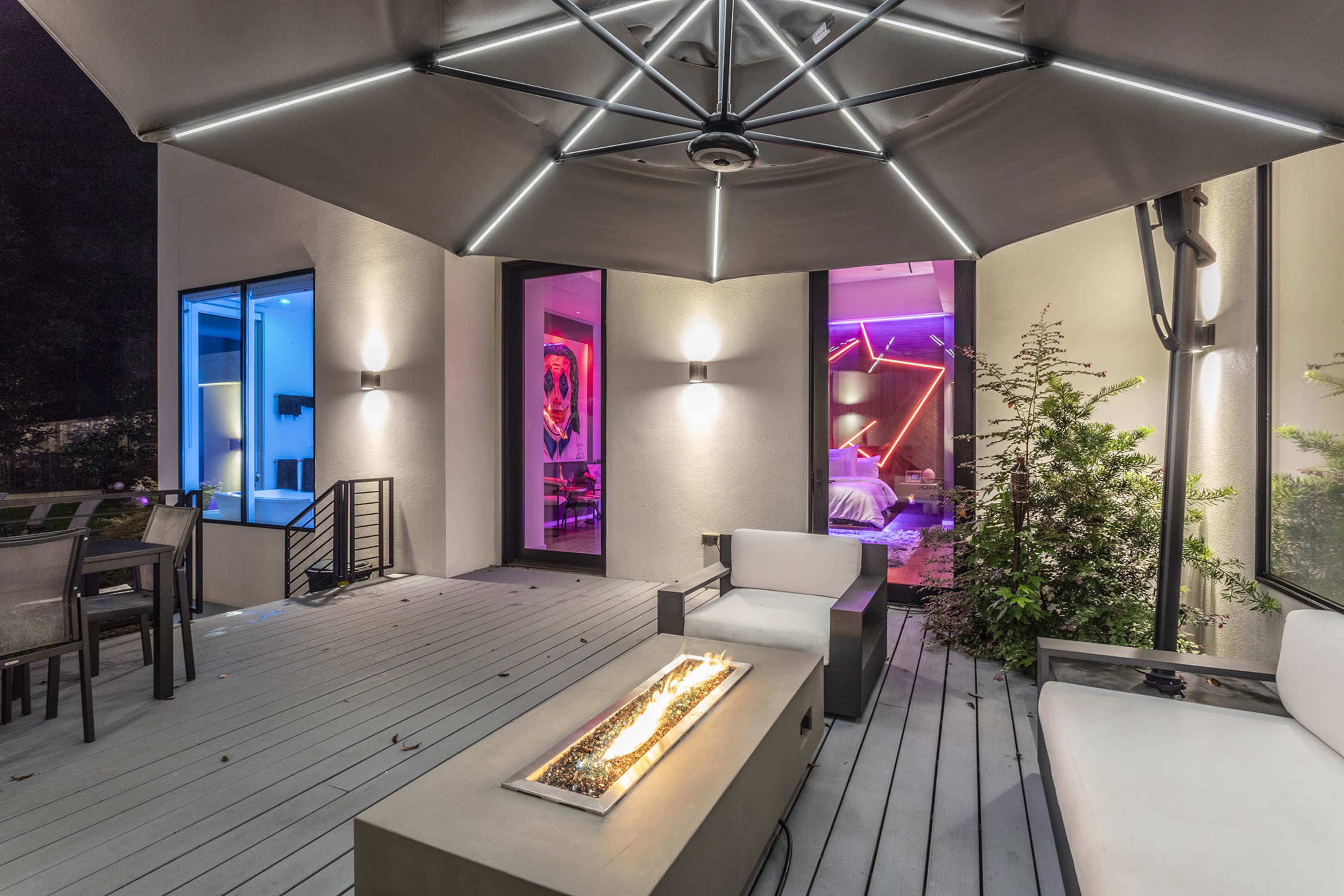 A modern outdoor seating area features a fire pit, sleek furniture, and an overhead canopy with integrated lighting, set against the backdrop of a stylish interior space.
