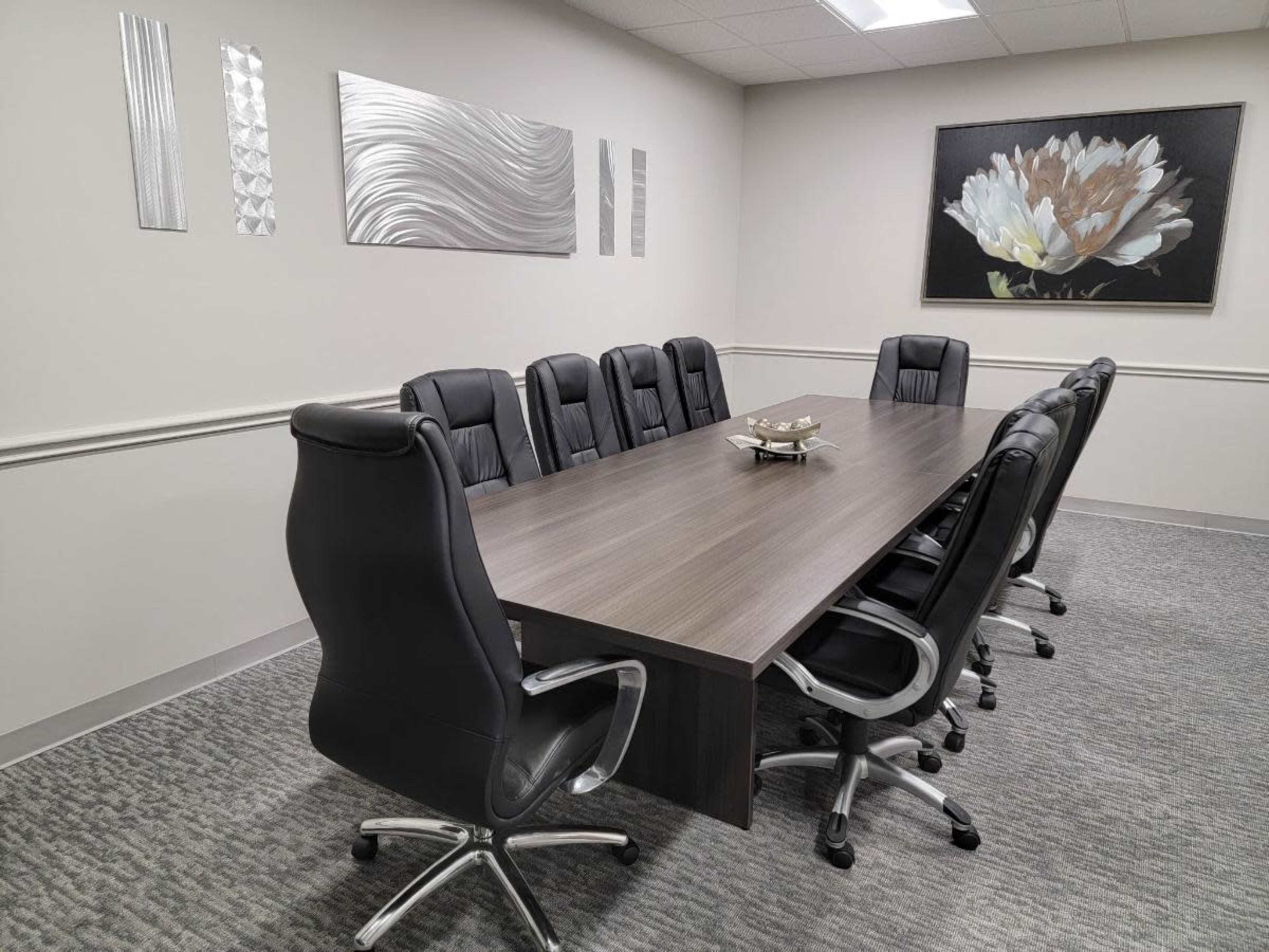 The image shows a conference room with a long rectangular table surrounded by black leather chairs and wall art featuring a flower.
