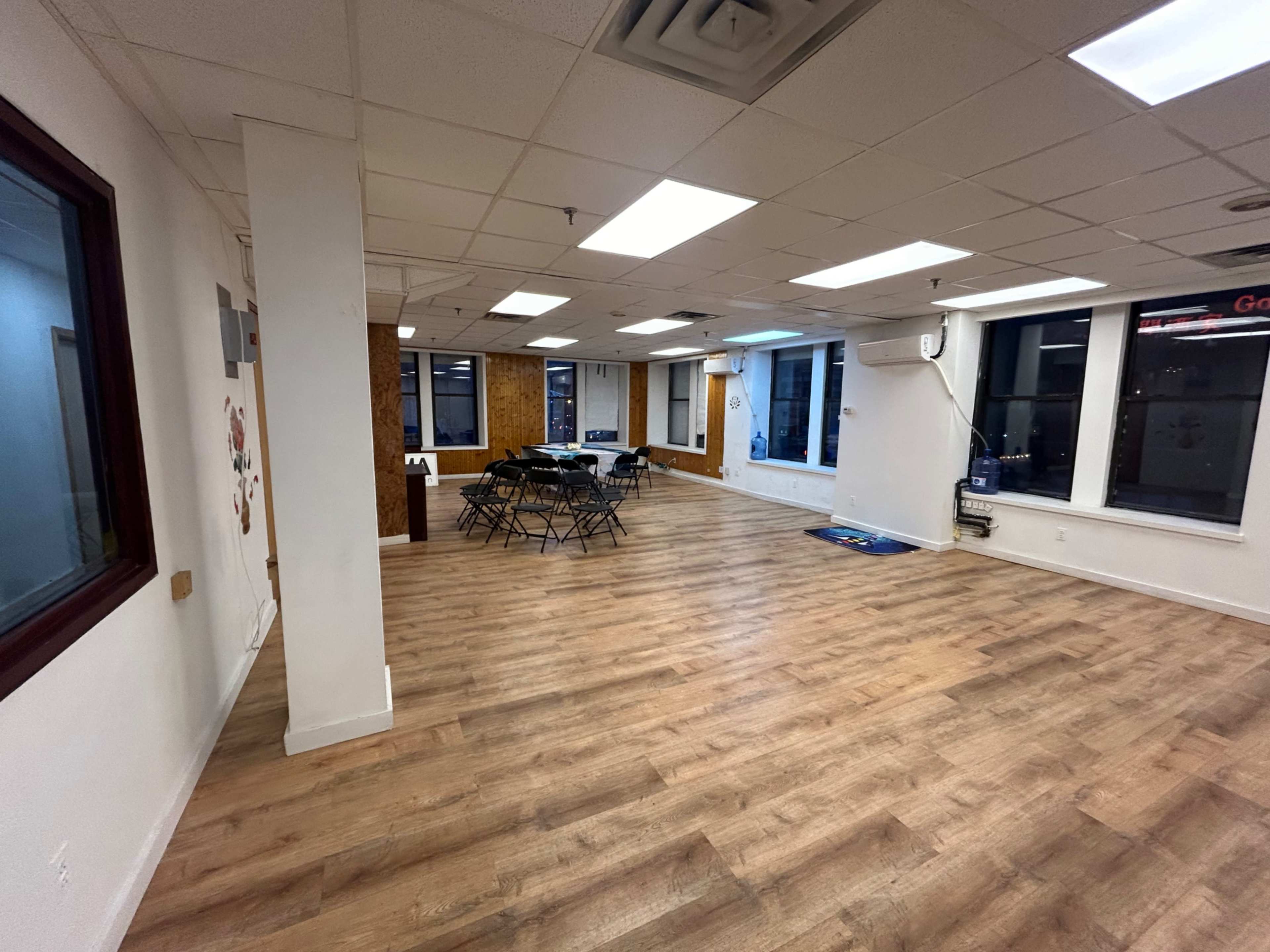 A spacious, empty room with wooden flooring, fluorescent lights, and a few chairs arranged near the walls.