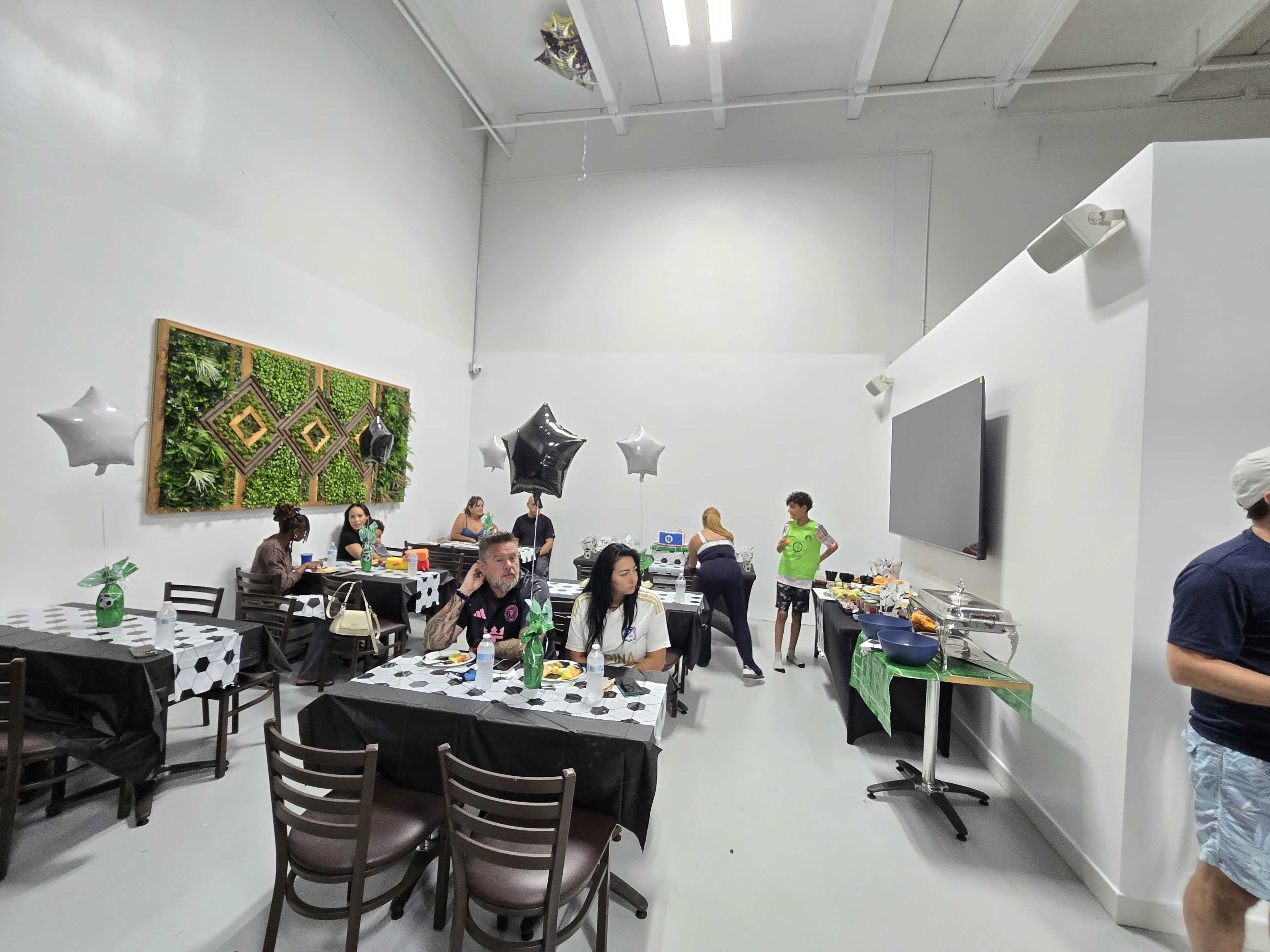 The image shows a gathering space with several tables decorated for a party, where people are interacting and food is set up on a side table.