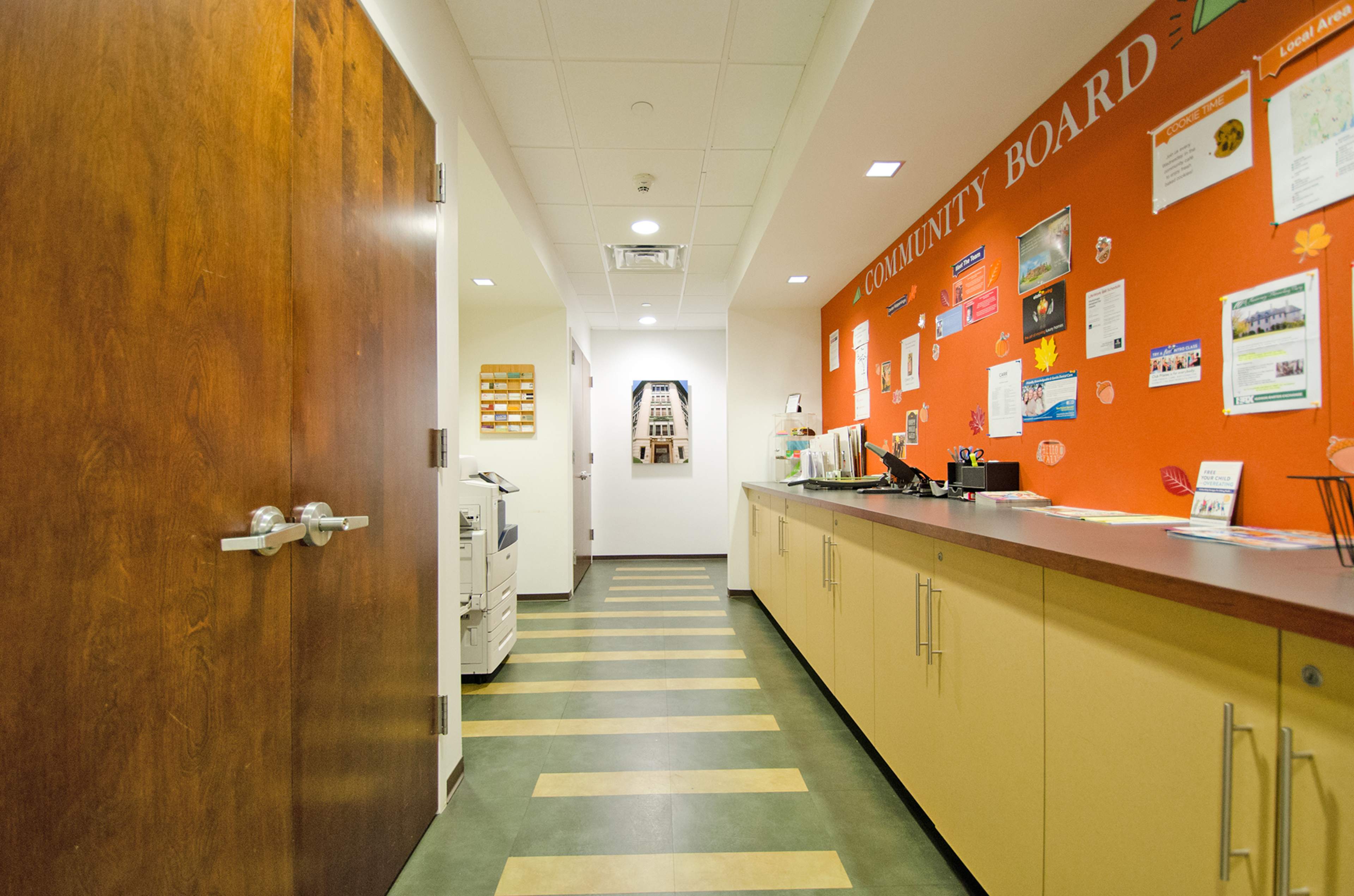 A well-lit hallway with a wooden door on the left and a long counter against an orange wall displaying a community board with various notices.