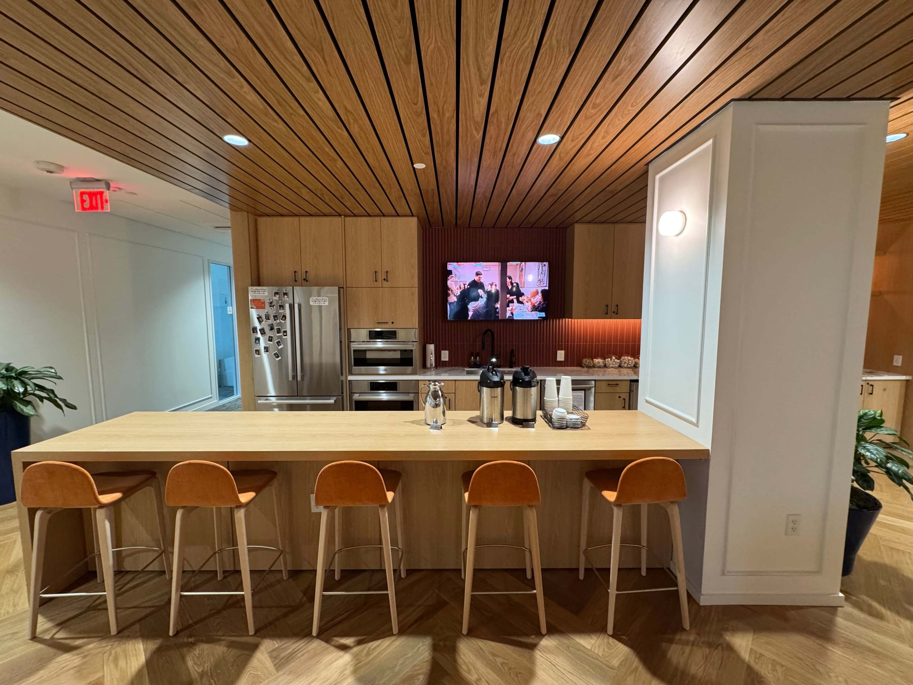 A modern kitchen area features a wooden bar with four stools, stainless steel appliances, and a television mounted on the wall.