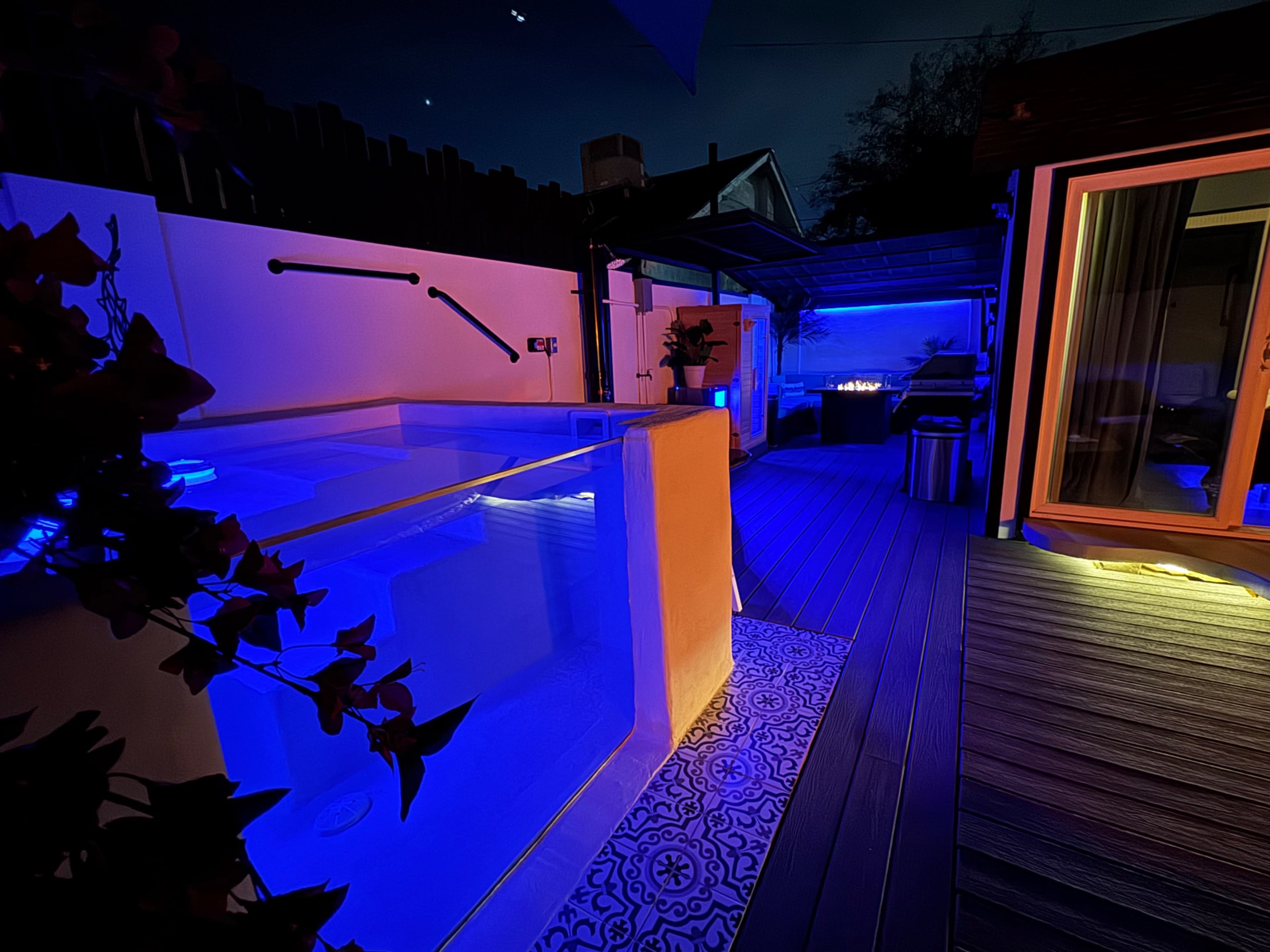 The image shows a modern outdoor space at night, featuring a spa illuminated with blue lights, surrounded by wooden decking and a cozy seating area.