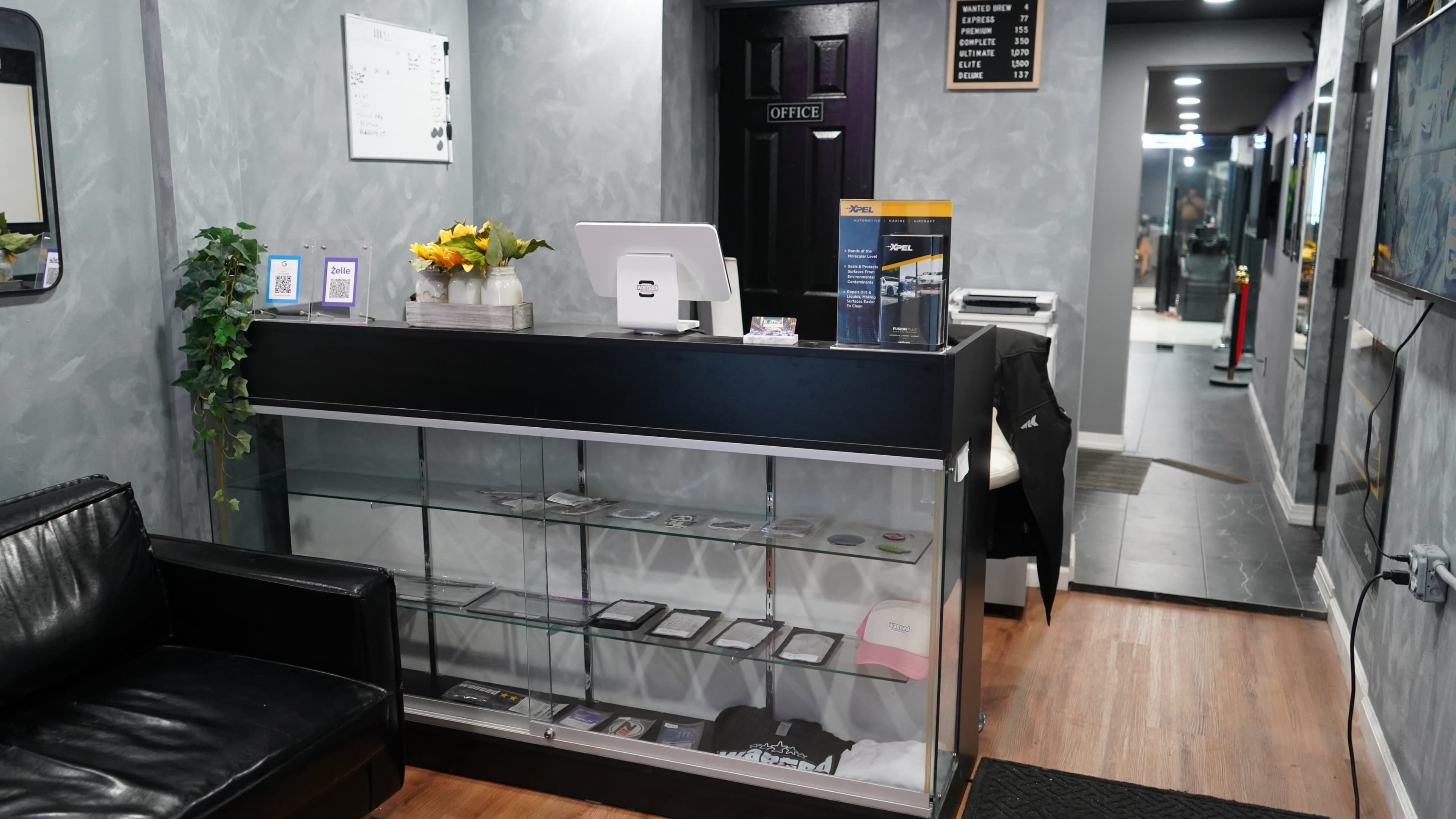 The image shows a modern reception area with a black front desk, glass display shelves, and a seating area featuring a black leather couch.