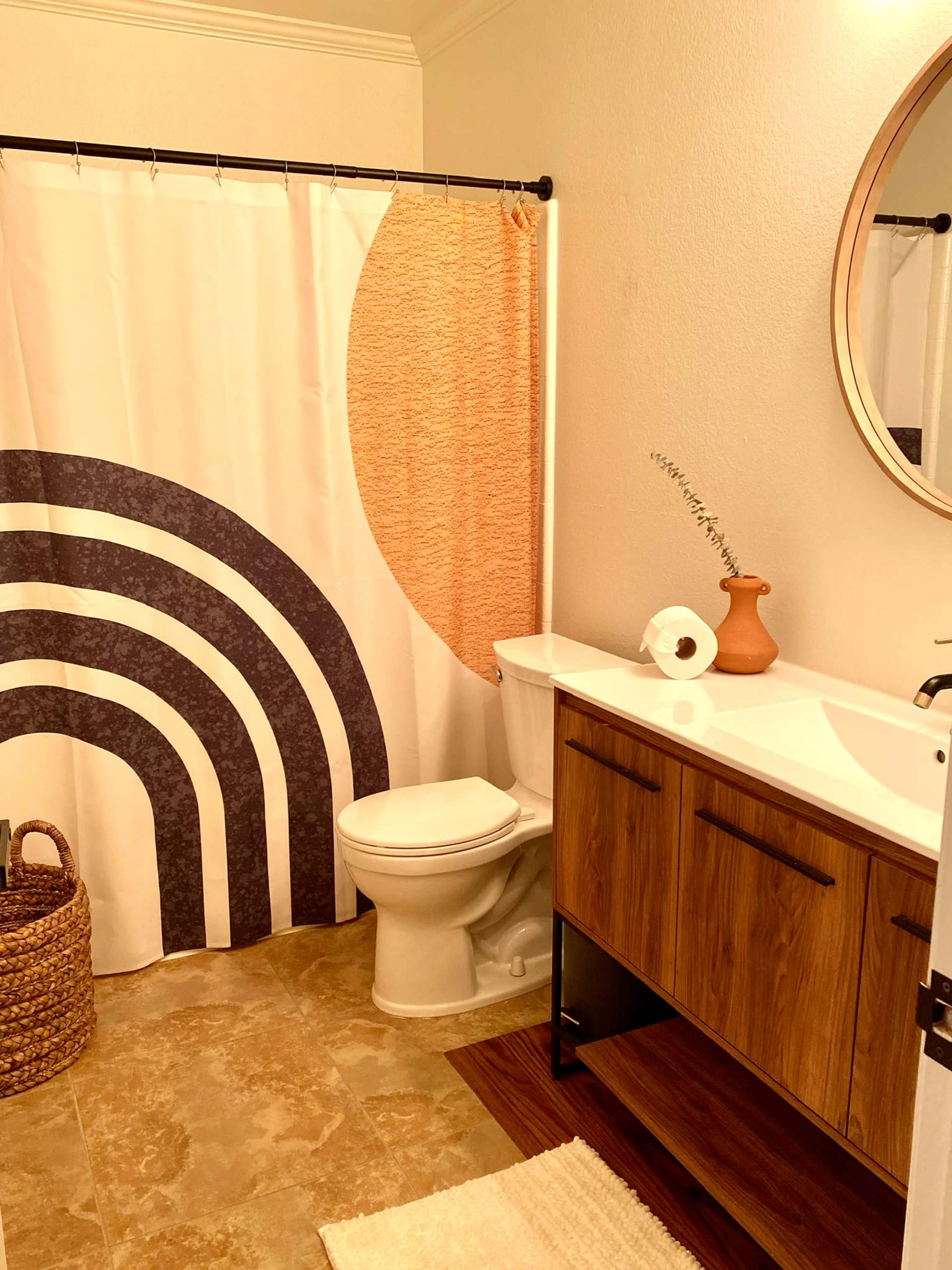 A bathroom featuring a shower curtain with a geometric design, a wooden vanity with a circular mirror, a toilet, and a woven basket in the corner.