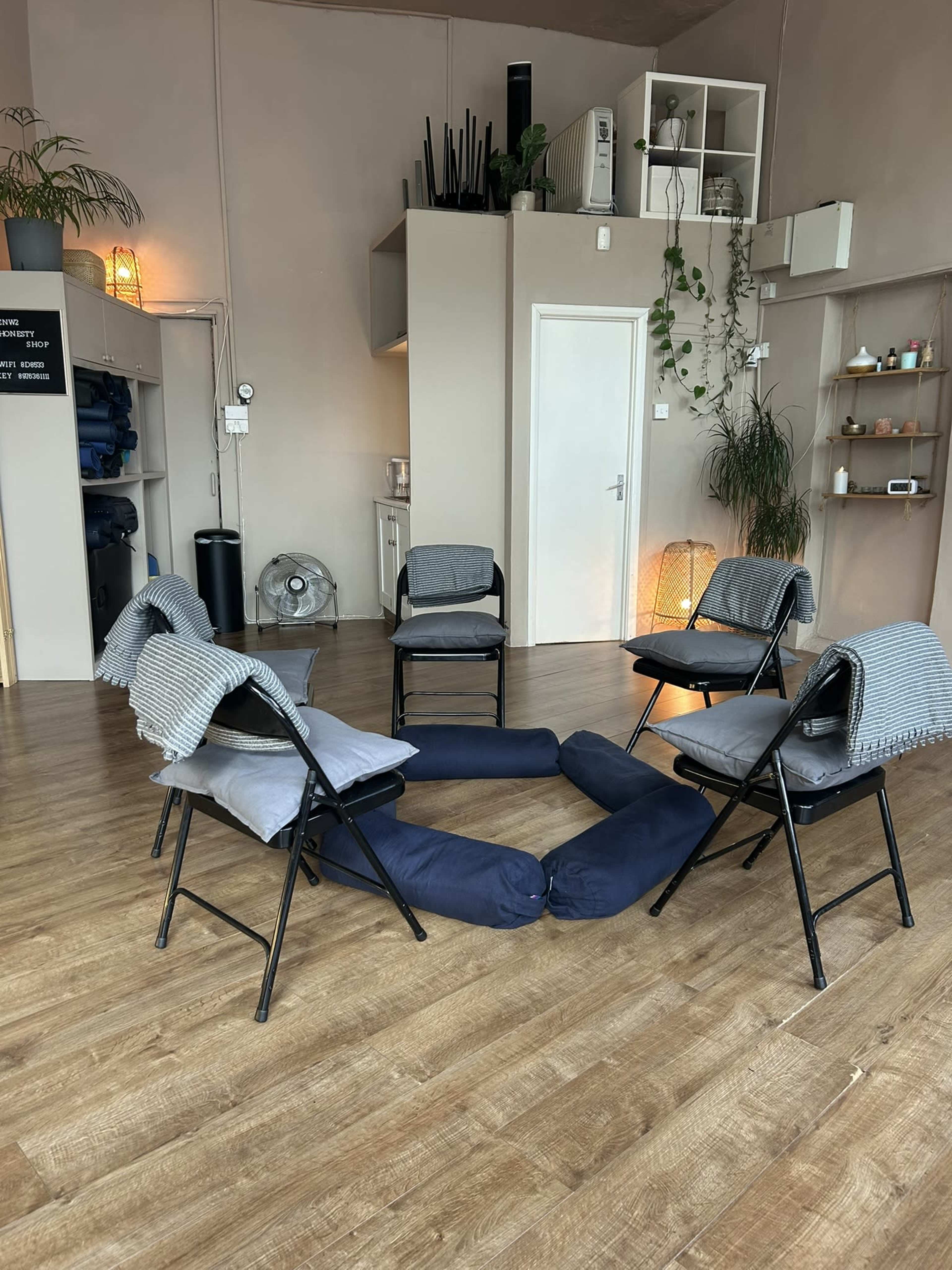 A circle of six folding chairs is arranged on a wooden floor, with two long cushions placed in the center and various plants and shelves visible in the background.
