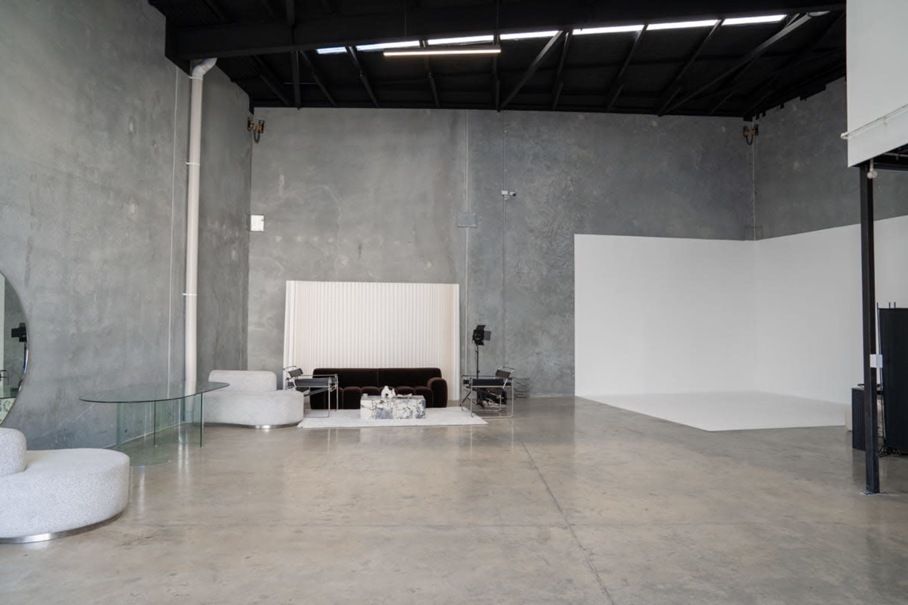 The image shows a minimalist studio space featuring concrete walls, a sofa, a glass table, and a large white backdrop area.