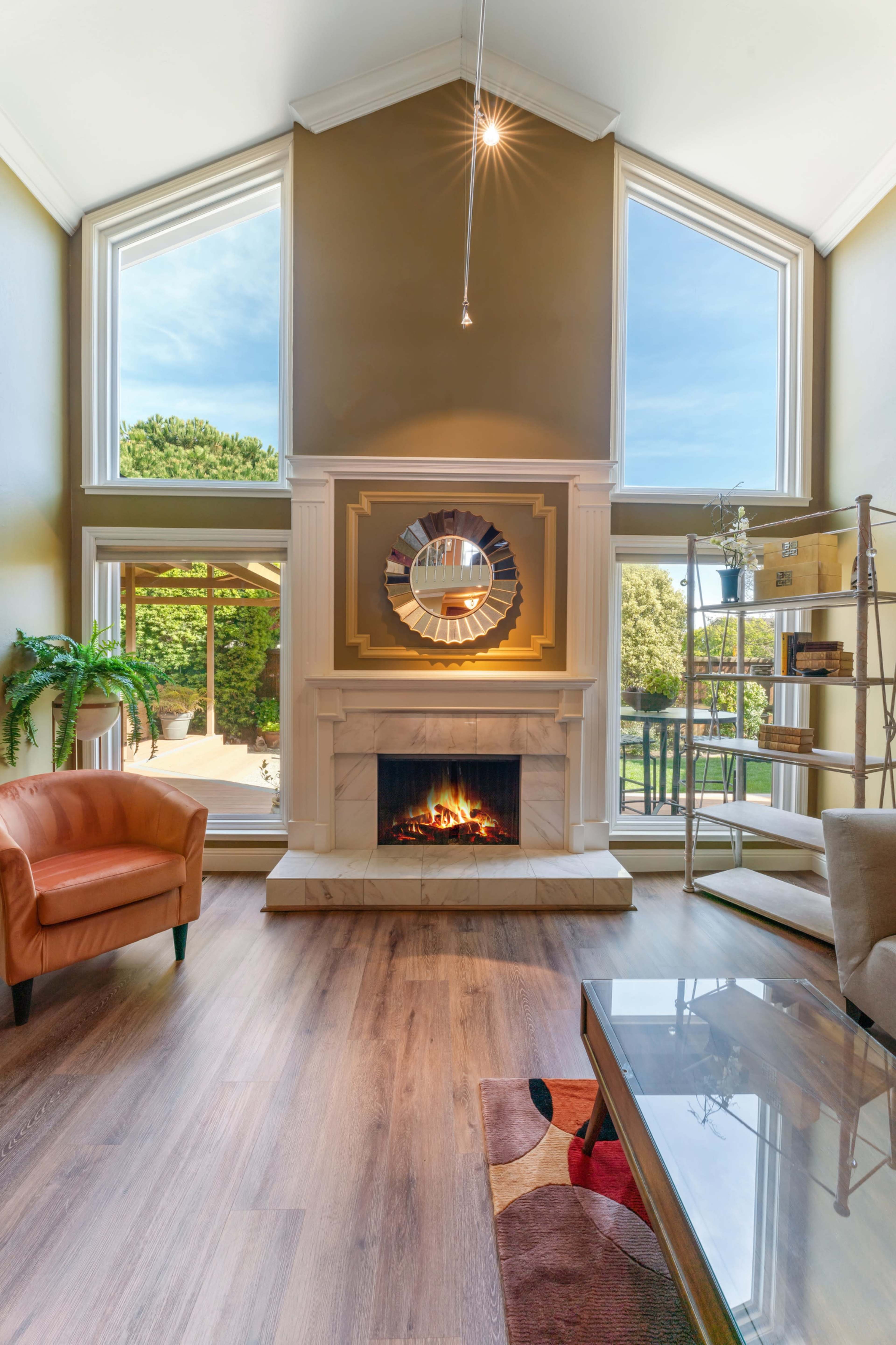 The living room features a fireplace centered beneath a large mirror, with tall windows allowing natural light and a view of the outdoors.