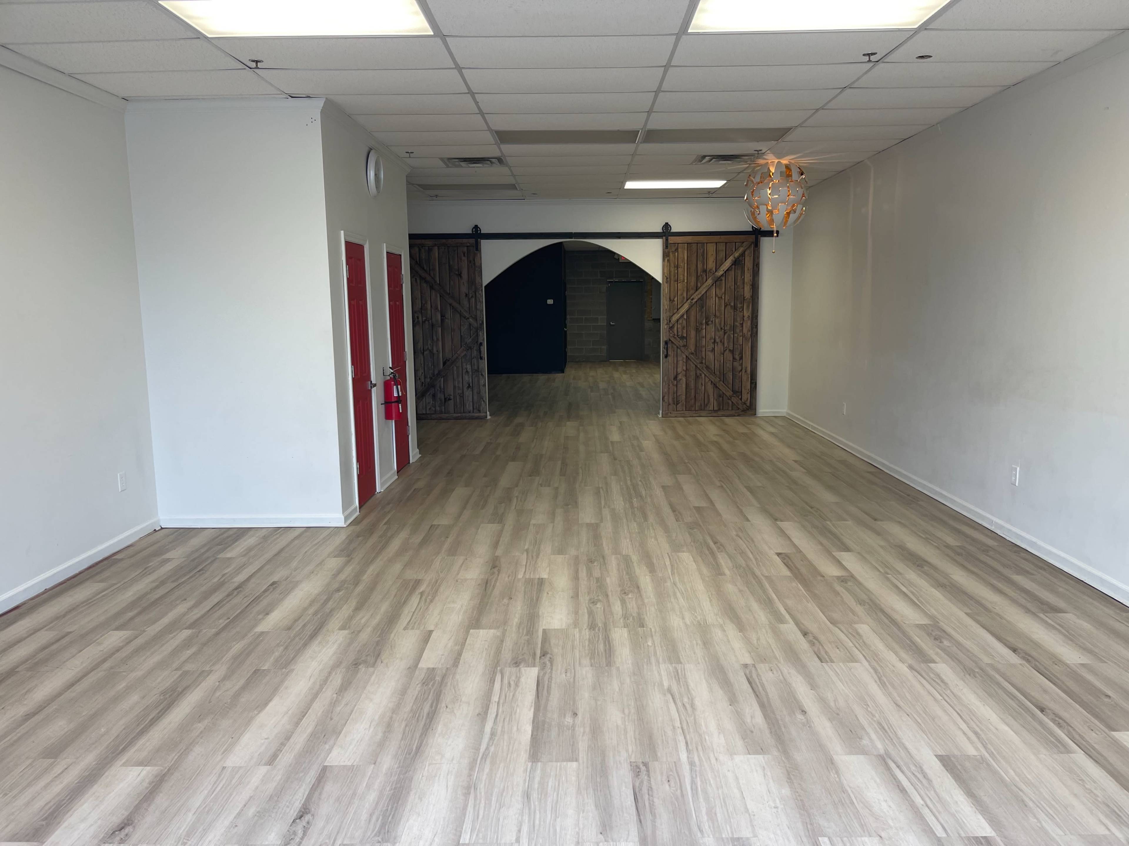 The image shows an empty retail space with wooden flooring, two large wooden doors at the back, and neutral-colored walls.